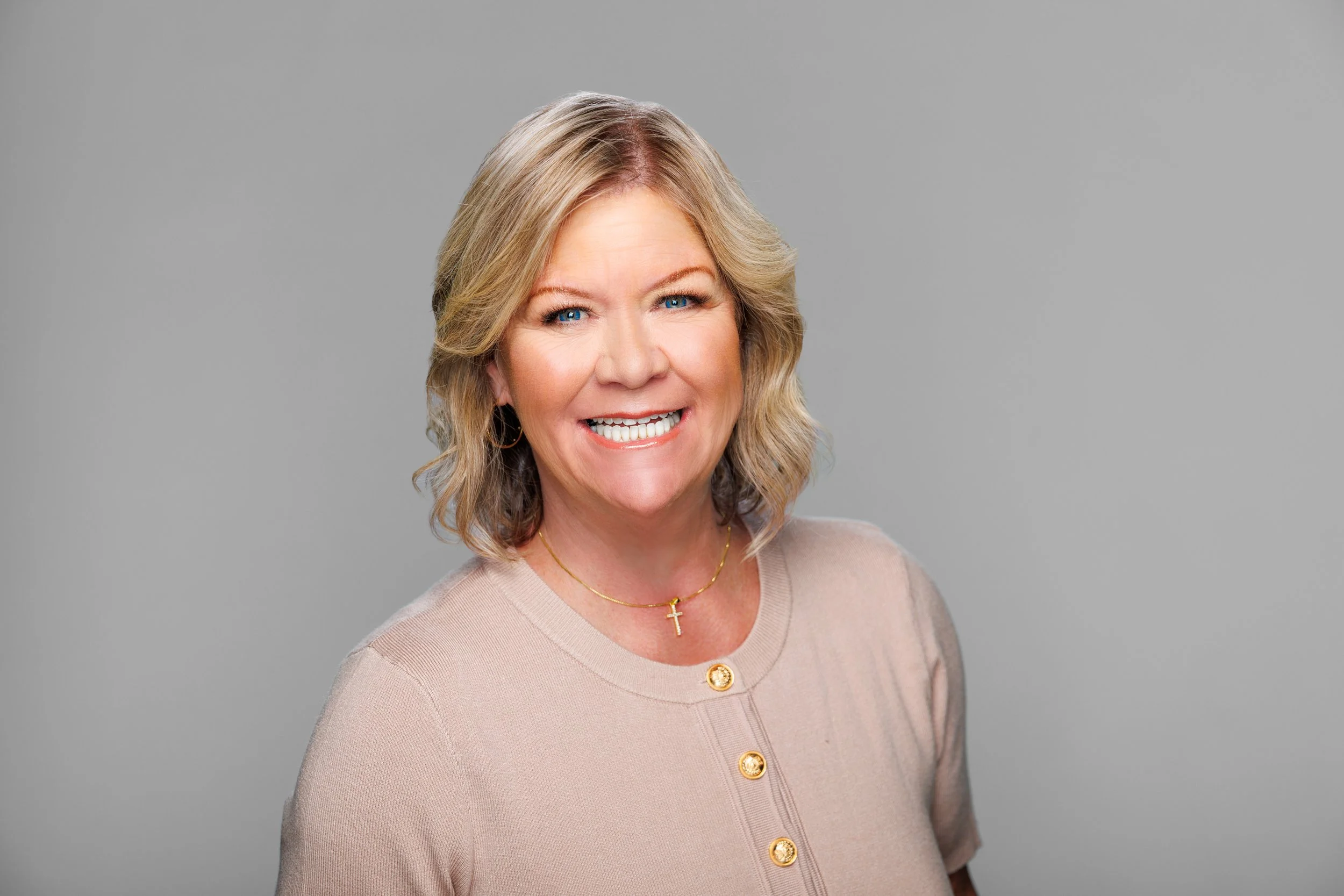 Portrait of a middle-aged woman with blonde hair, blue eyes, smiling, wearing a beige top, gold necklace with a cross pendant, and gold buttons, against a plain gray background.