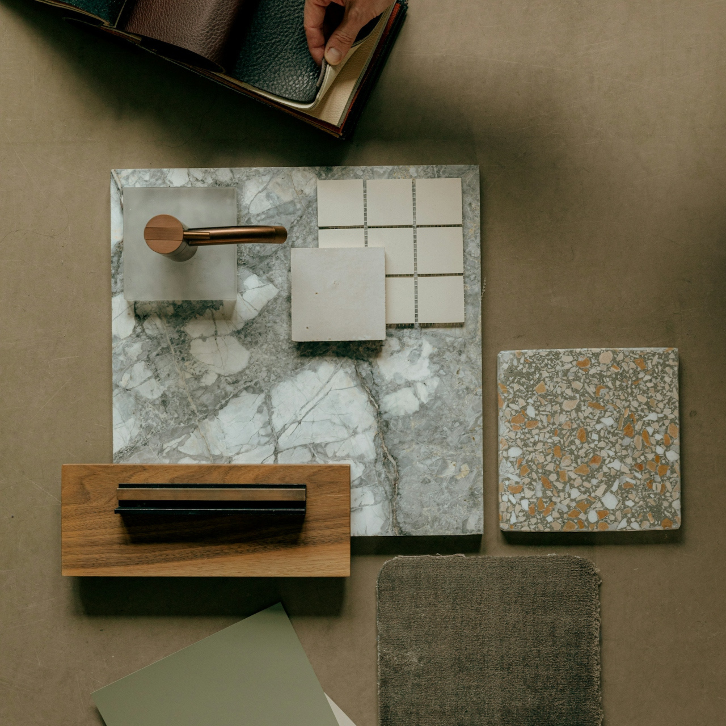 Arrangement of various tile samples on a marble slab surface, including different colors and textures such as ceramic, stone, and terrazzo, along with color swatches and a copper-colored fixture.