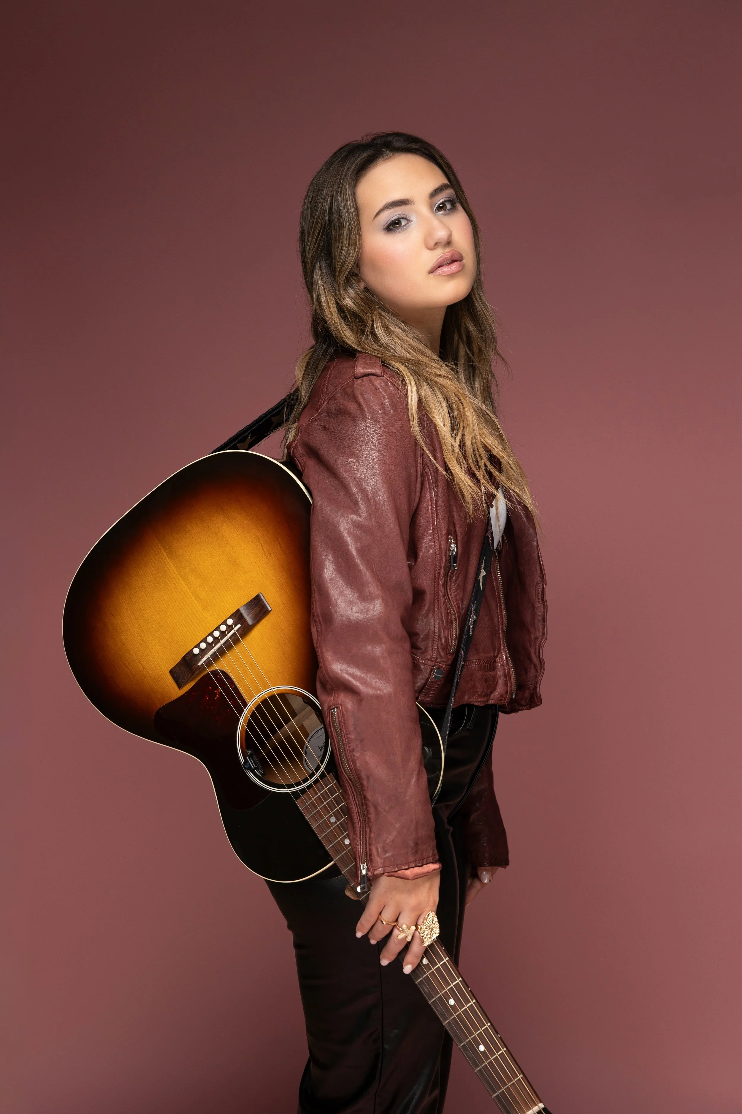 A young woman with long brown hair and a leather jacket, holding an acoustic guitar, standing against a mauve background.