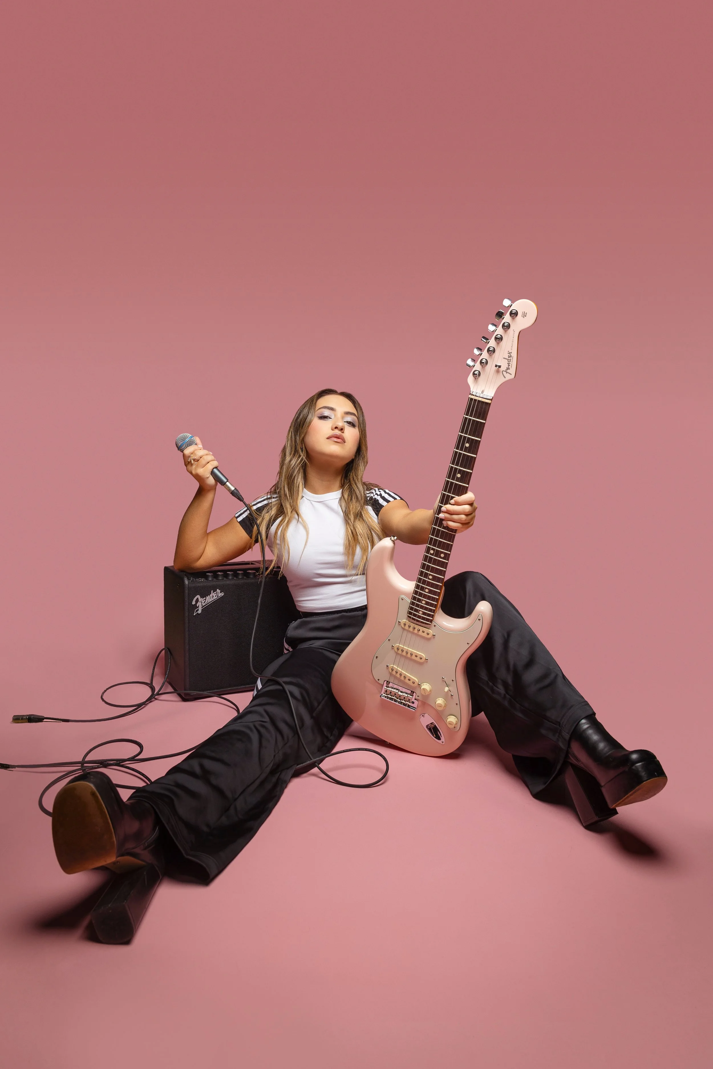 Young woman sitting on the floor against a pink background, holding a pink electric guitar and a microphone, with an amplifier nearby, dressed in a white t-shirt with black Adidas stripes and black pants.