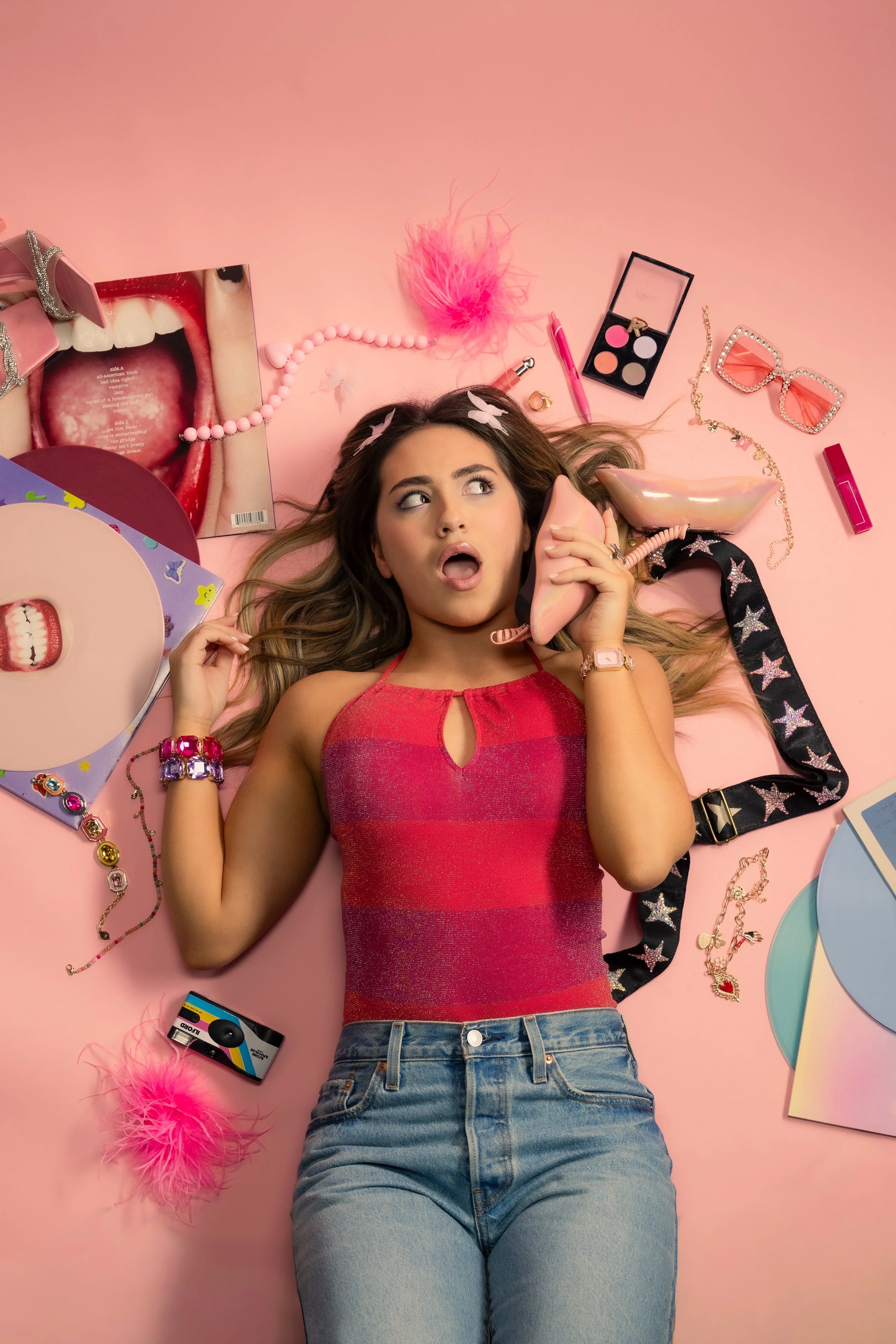 Woman lying on pink background surrounded by makeup and accessories, talking on a pink rotary phone.