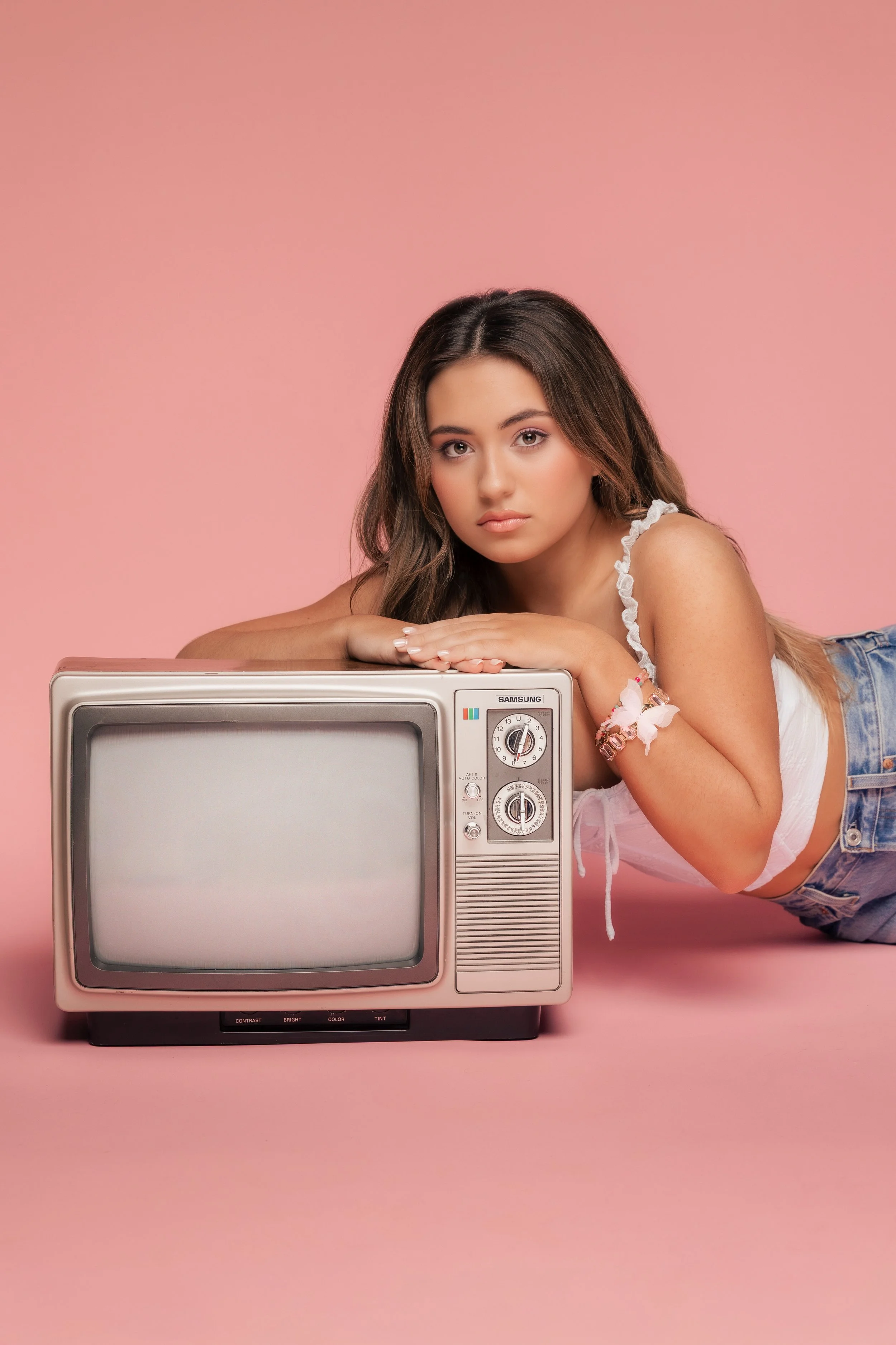 A young woman with wavy brown hair and wearing a white ruffled tank top and denim shorts is lying on her stomach on a pink background. She is resting her chin on her folded arms and looking at the camera. In front of her is a vintage pink and gray television set with control dials and a small screen.