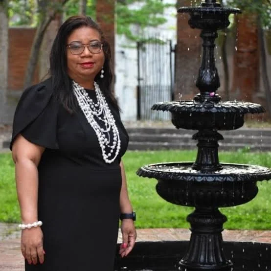 A woman wearing glasses and a black dress with pearl jewelry stands next to a black outdoor fountain in a backyard with green grass and trees.