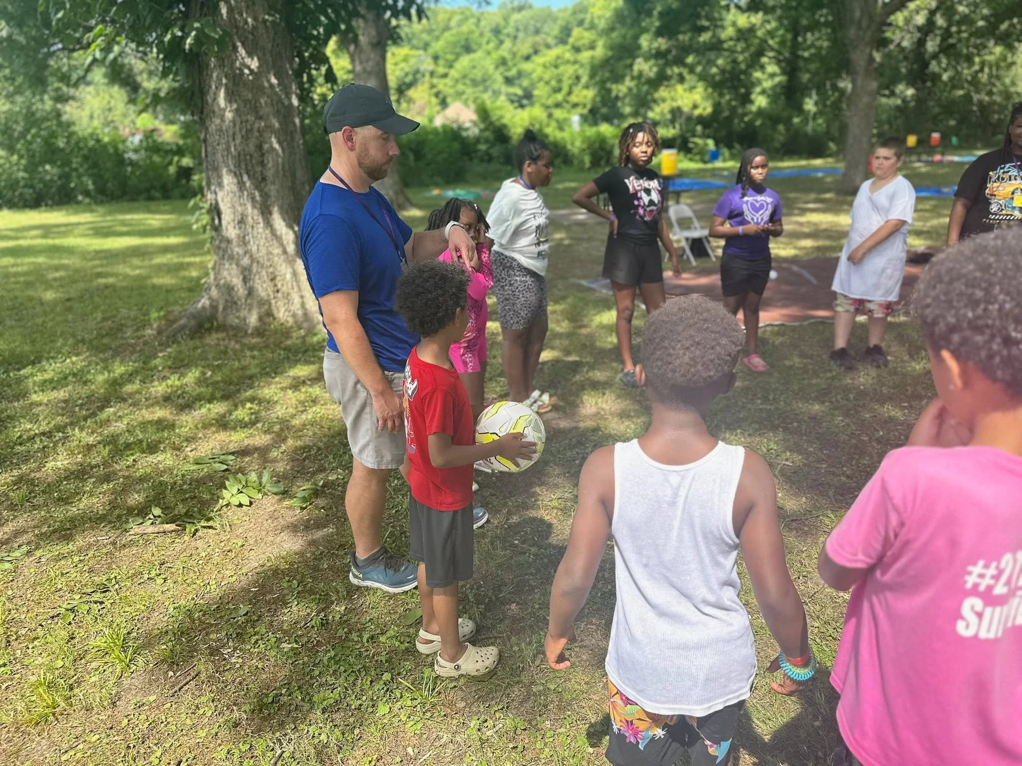 A group of children and adults standing outdoors under a large tree in a grassy area, possibly participating in a group activity or gathering on a sunny day.