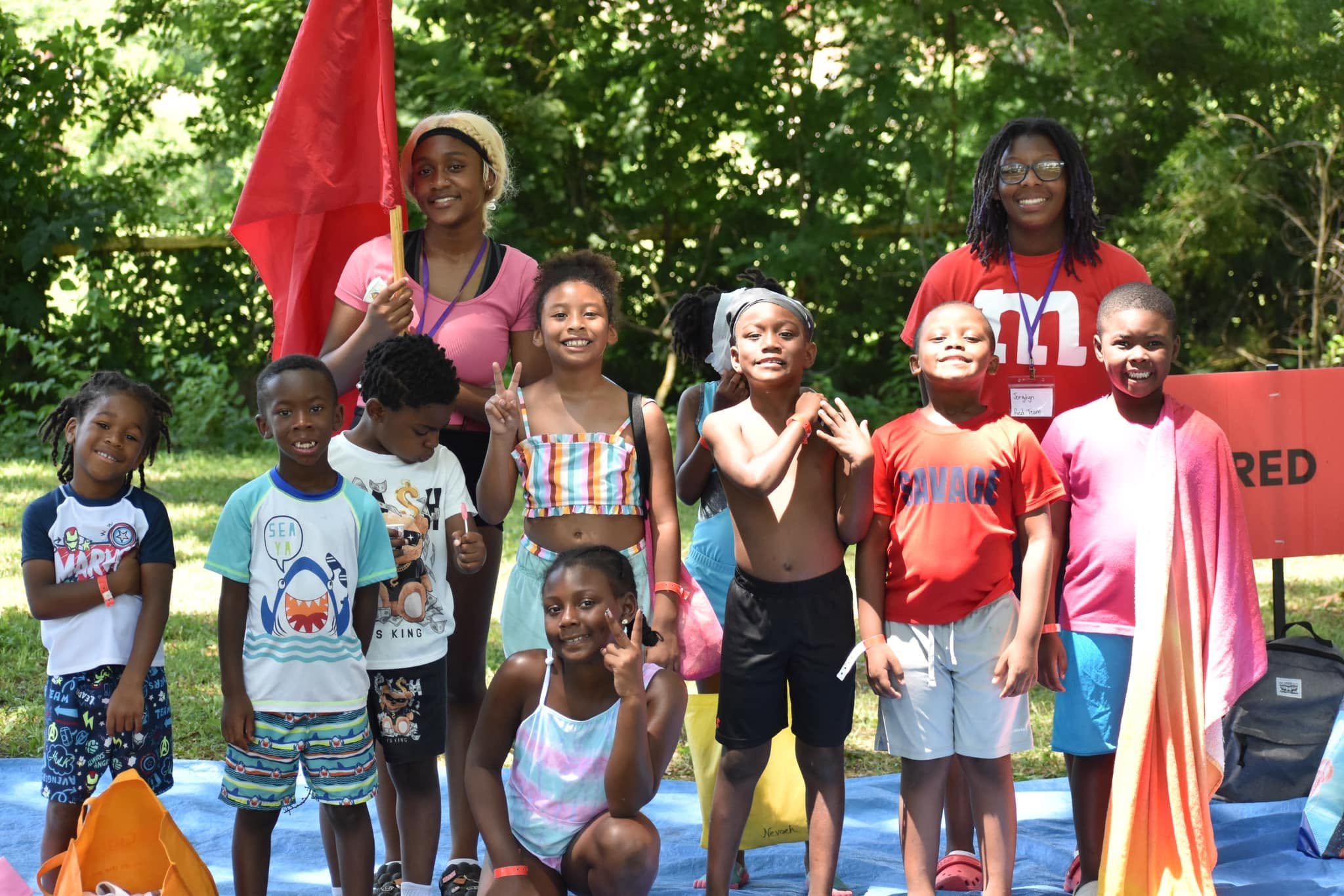 A group of children and two women standing outdoors in front of trees, smiling for a group photo. The children are wearing colorful casual clothing, some making peace signs, others with arms crossed or hands on hips. One woman is holding a red flag, 