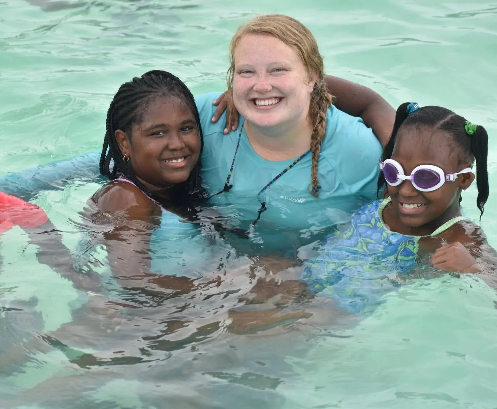 A group of three people, including a woman with red hair and two young girls, in a swimming pool smiling and enjoying their time together.