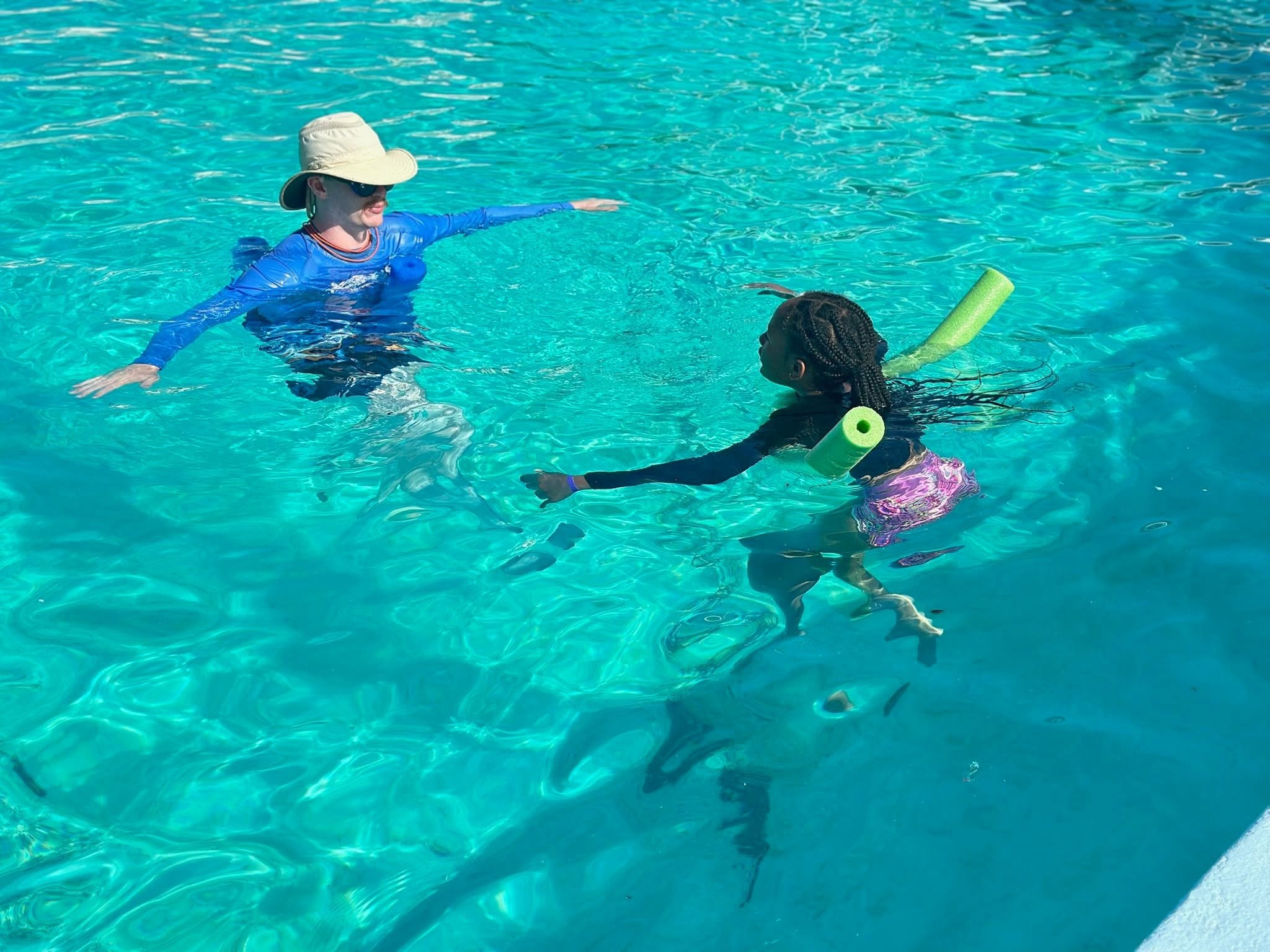 Two people swimming in a clear blue swimming pool, one wearing a wide-brimmed hat and sunglasses, the other with braided hair and a pool noodle.