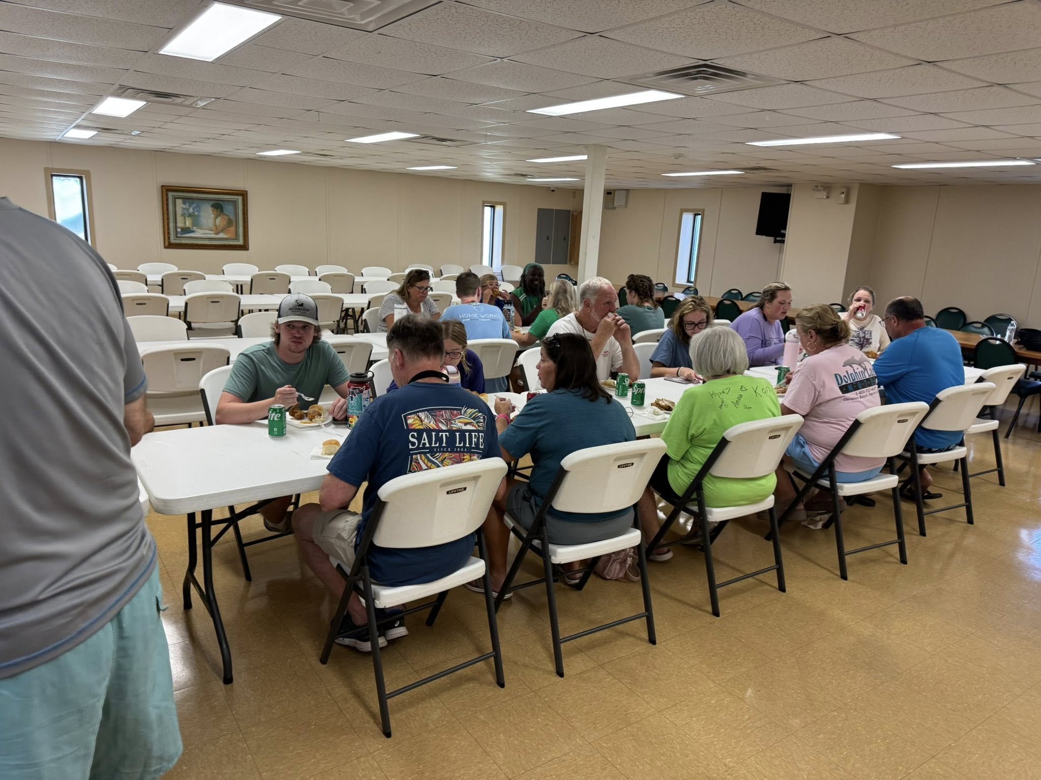 People sitting at tables having a meal in a large room with beige walls, ceiling tiles, and small windows.