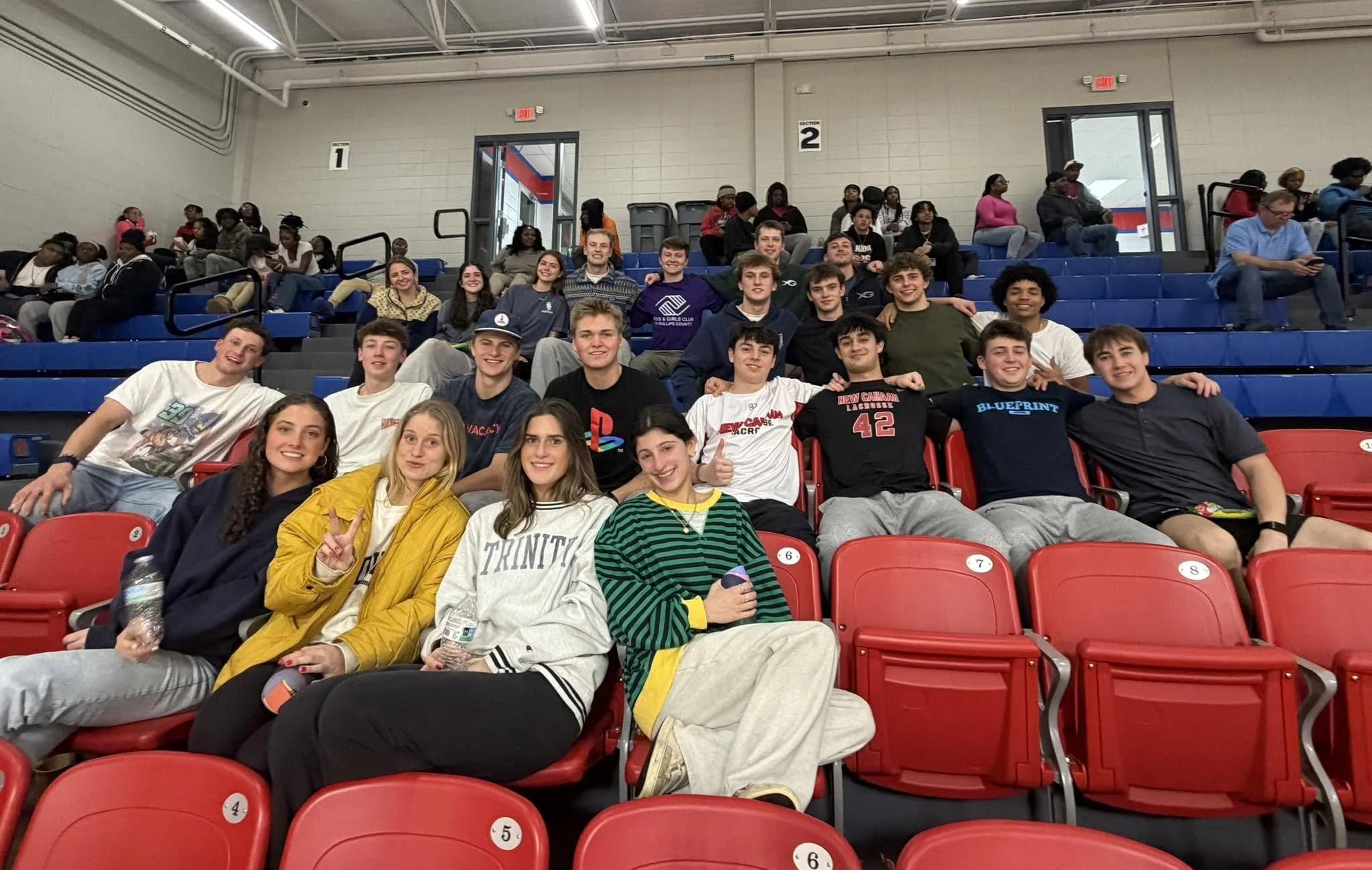 Group of young people sitting on red and blue bleachers inside a gymnasium or auditorium, smiling and posing for the photo.