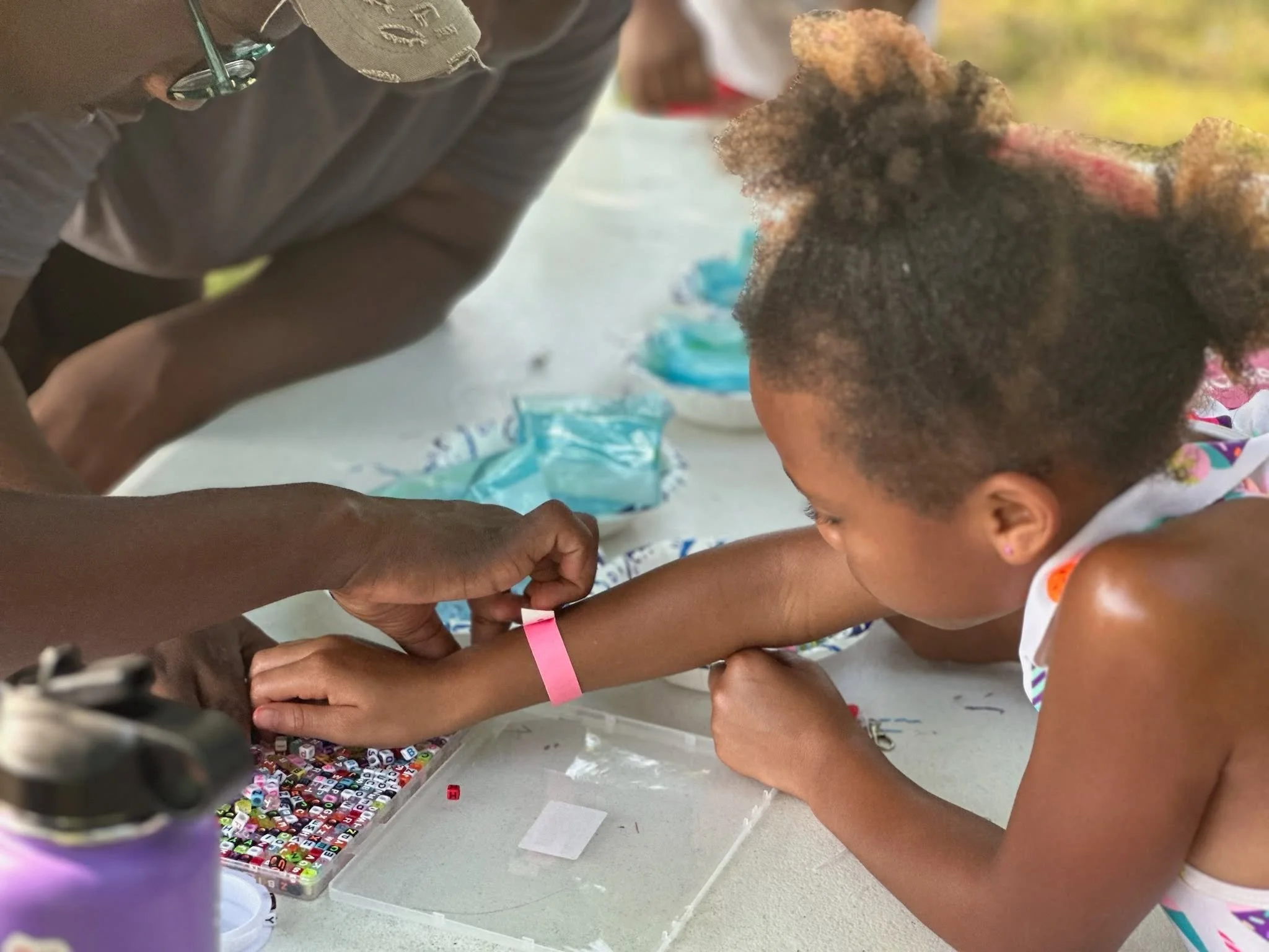 Child and adult working together on a craft project with colorful beads, with the child's hair styled in a curly afro, and the child's arm has a pink wristband.