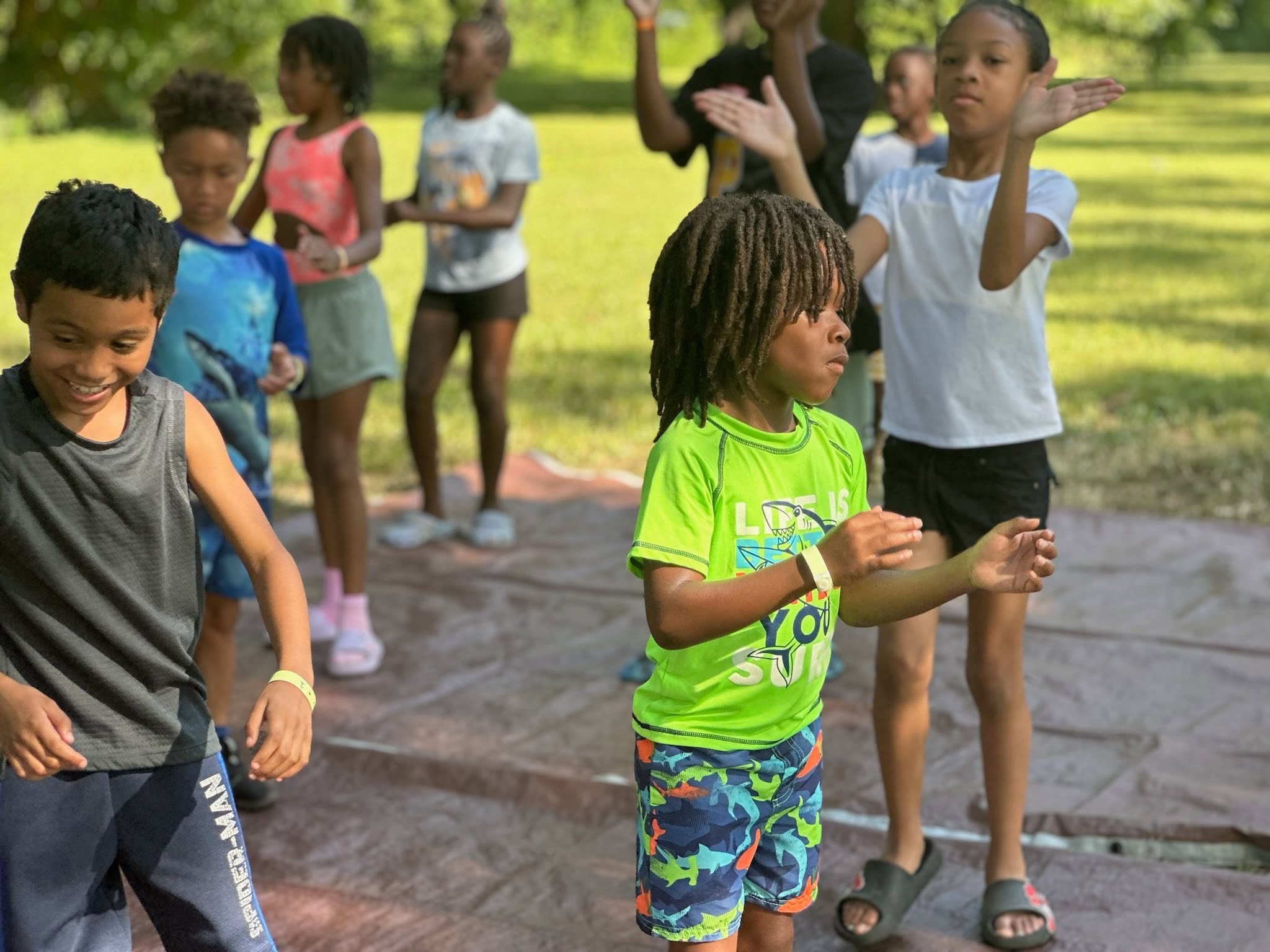 A group of children playing outdoors in a park with trees in the background. They are engaged in an activity, and some are clapping while others watch.