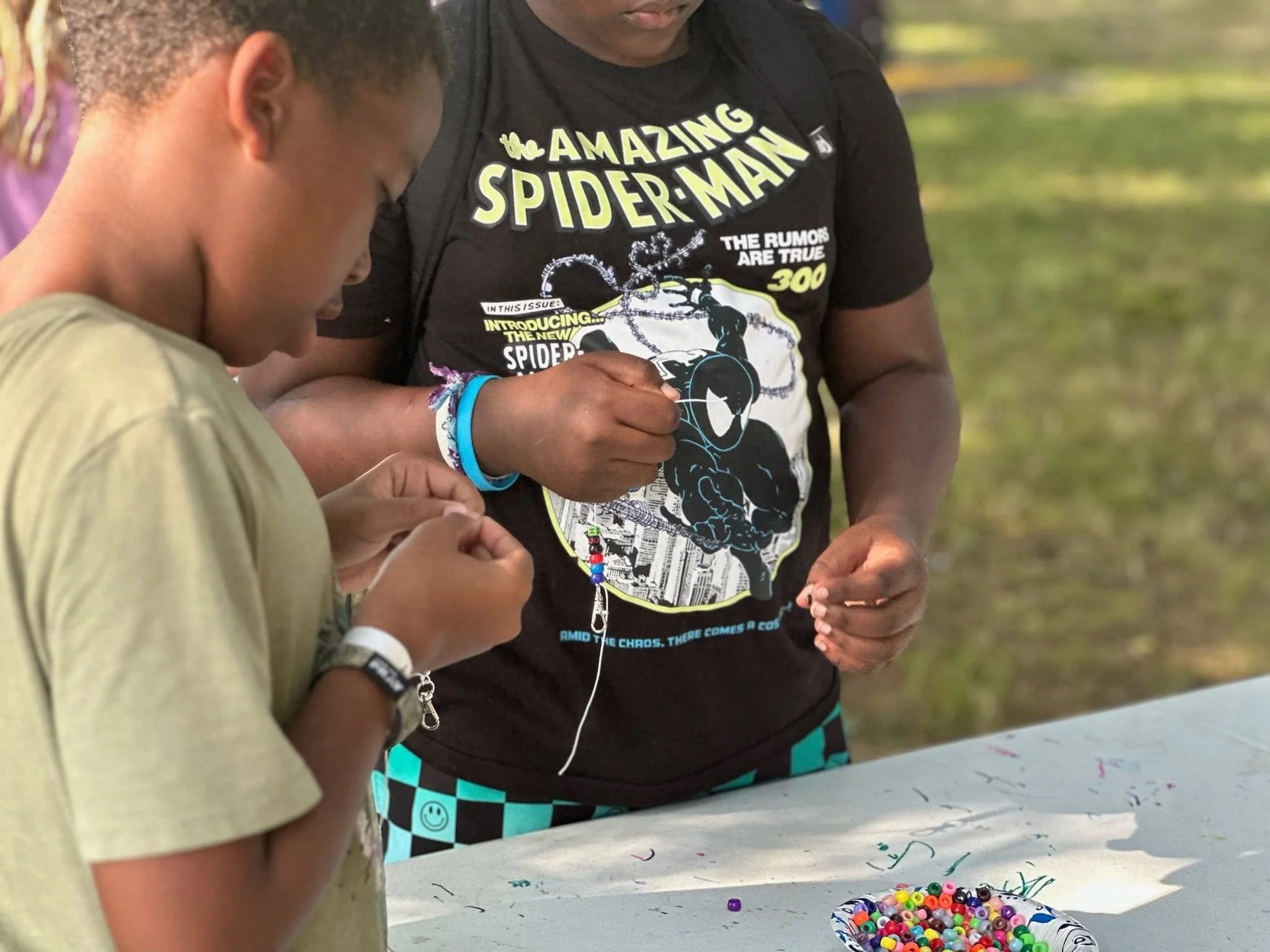 A young boy and a man are decorating objects with colorful beads at a table outdoors. The boy is wearing a beige shirt and a bracelet, and the man is wearing a black Spider-Man t-shirt.