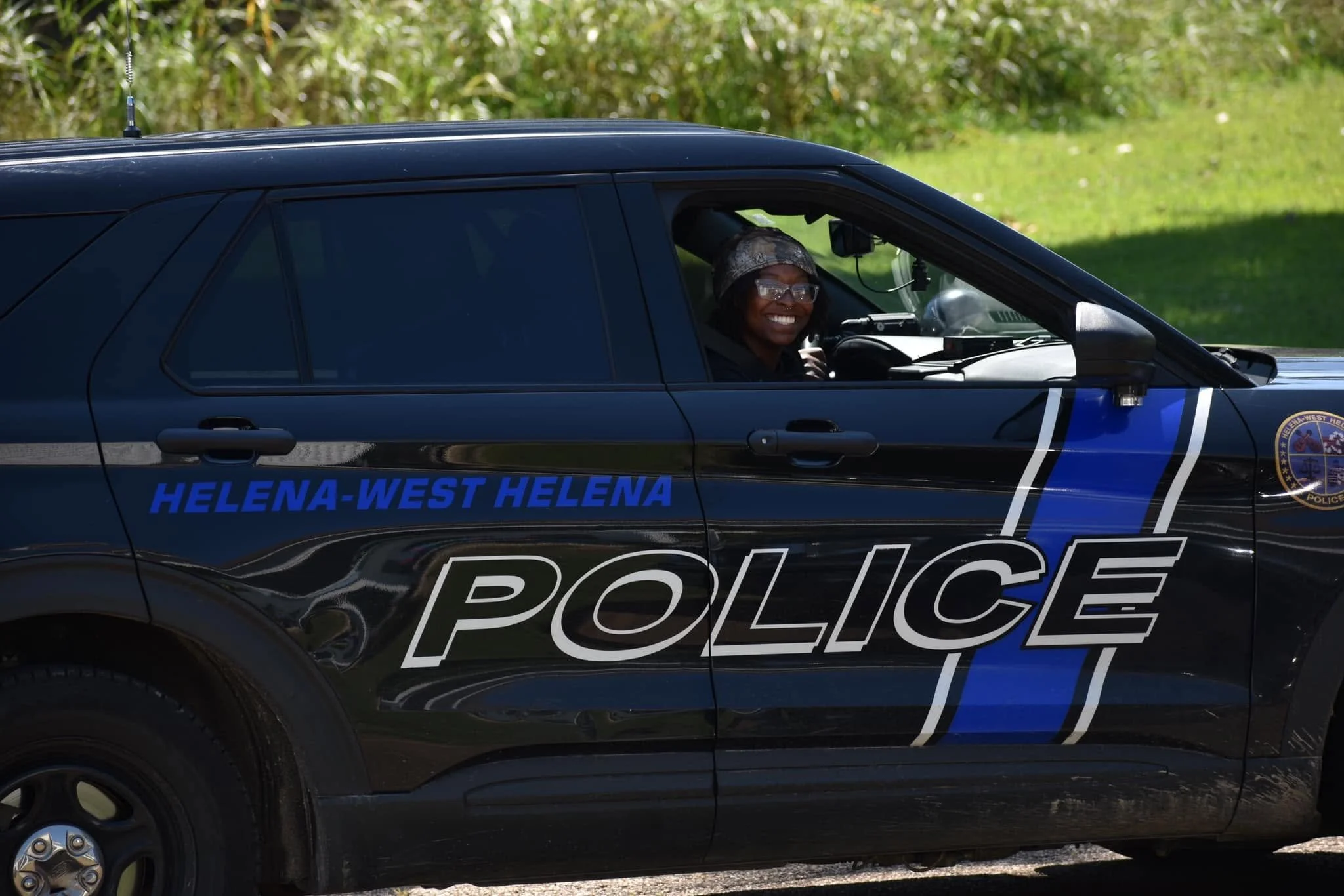 A smiling female police officer with glasses sitting in the driver's seat of a Helena-West Helena police car, with a grassy field and some trees in the background.