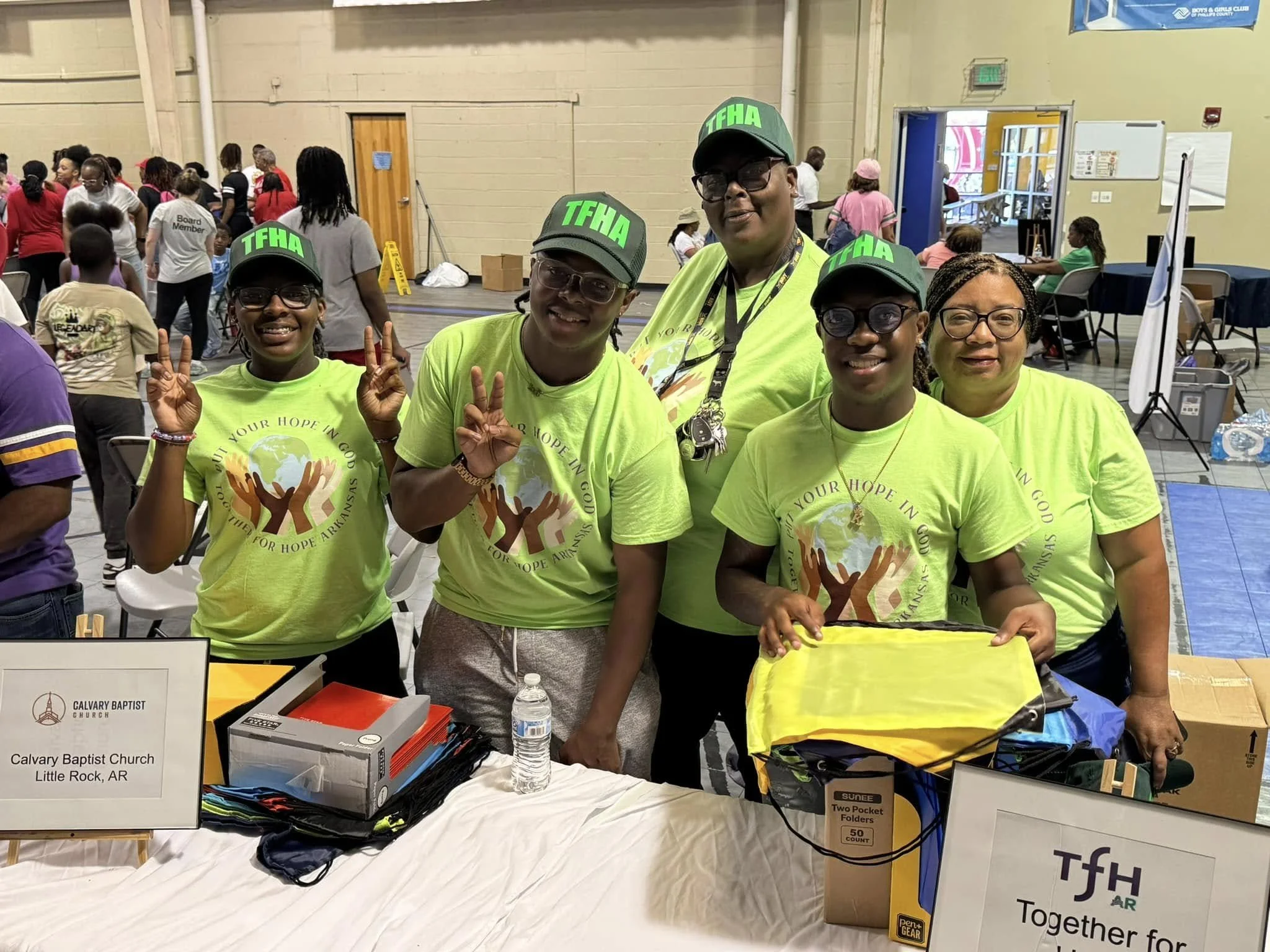 Five smiling women wearing bright yellow T-shirts and green caps with 'TFHA' written in green. They are standing behind a table at an indoor event, with other people in the background. The table displays various items including a water bottle, folders, and a sign indicating 'Calvary Baptist Church Little Rock AR.'