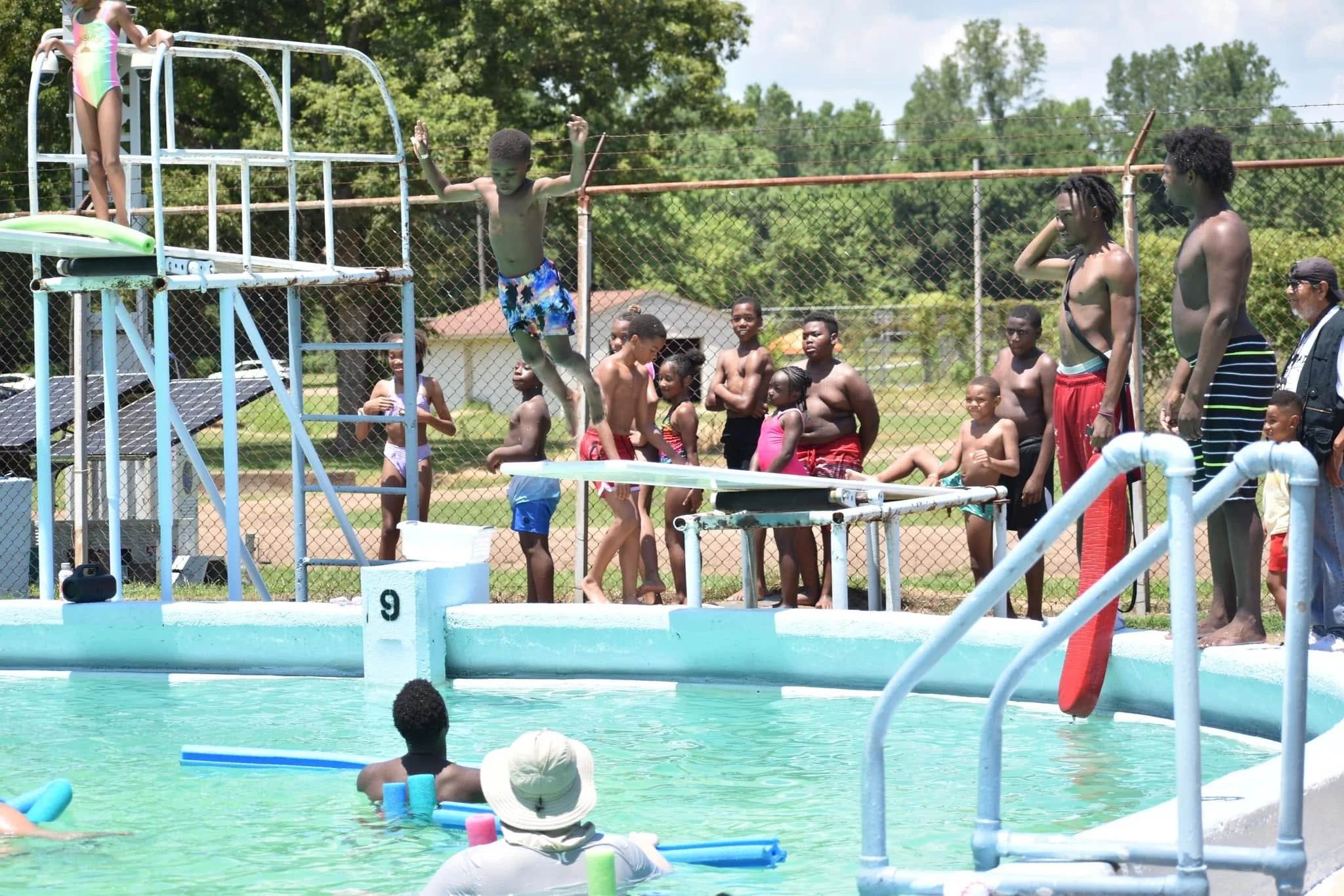 Children and adults enjoying a swimming pool and water slide during a sunny daytime, with trees and a fence in the background.