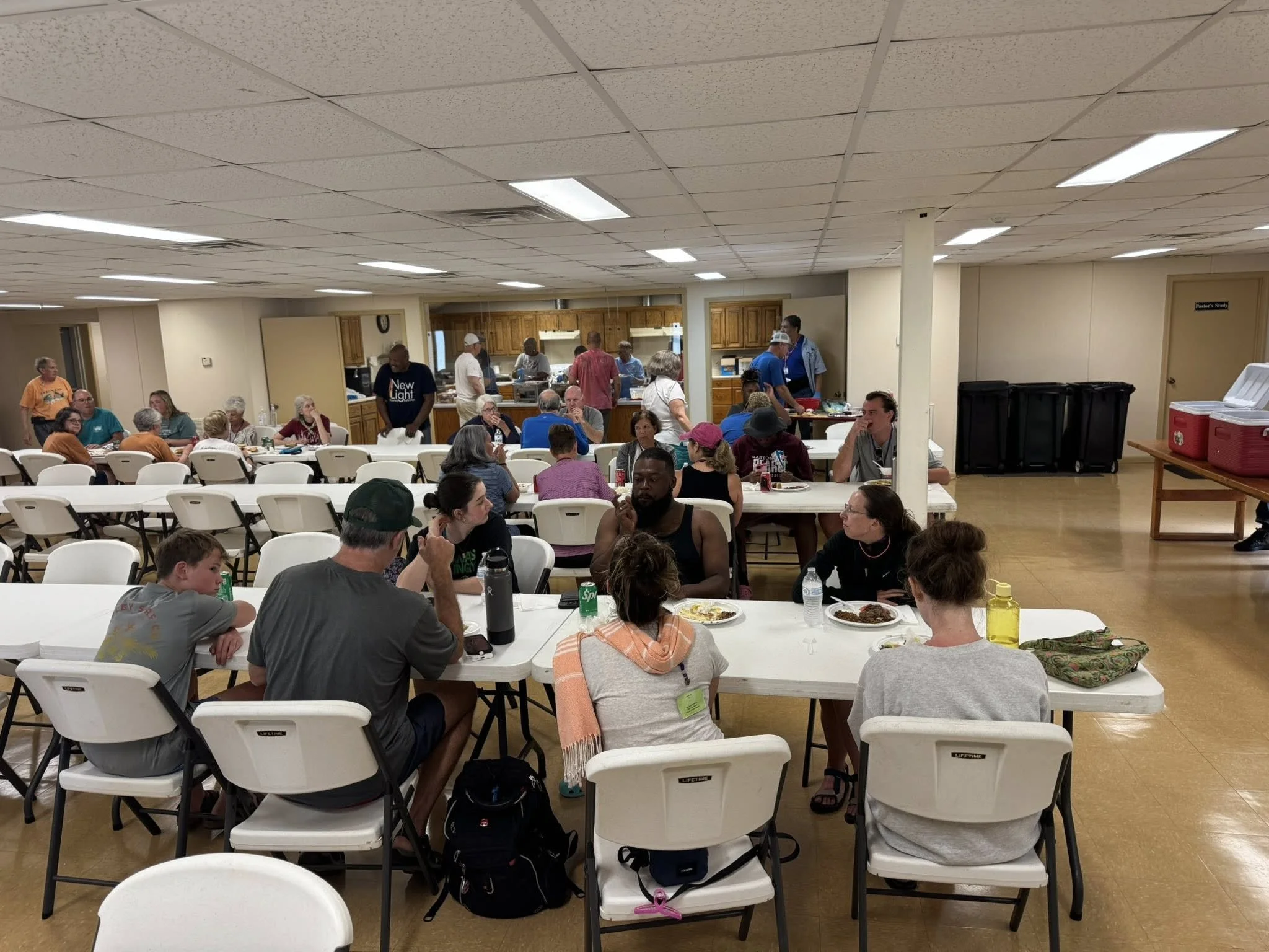 A large indoor gathering with people sitting at white folding tables and chairs, eating and socializing, with a kitchen area in the background and some people standing.