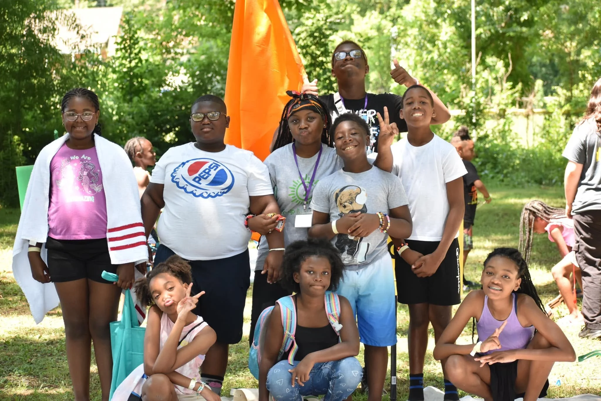 Group of children and adults outdoors, smiling, posing for a photo at a park or outdoor event, some making peace signs, with lush green trees in the background.
