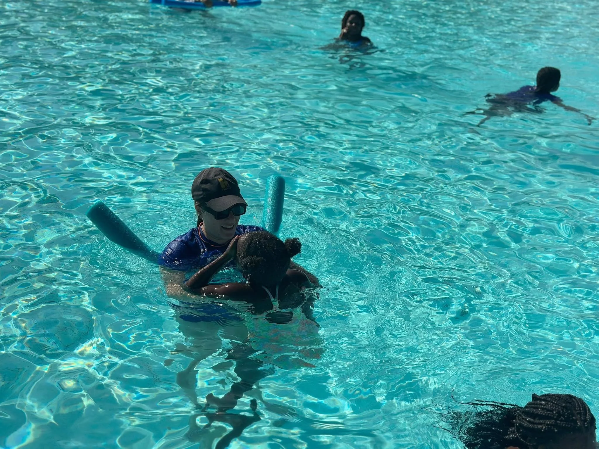 A person with a hat and sunglasses holding a child in a swimming pool, with two other children swimming in the background.