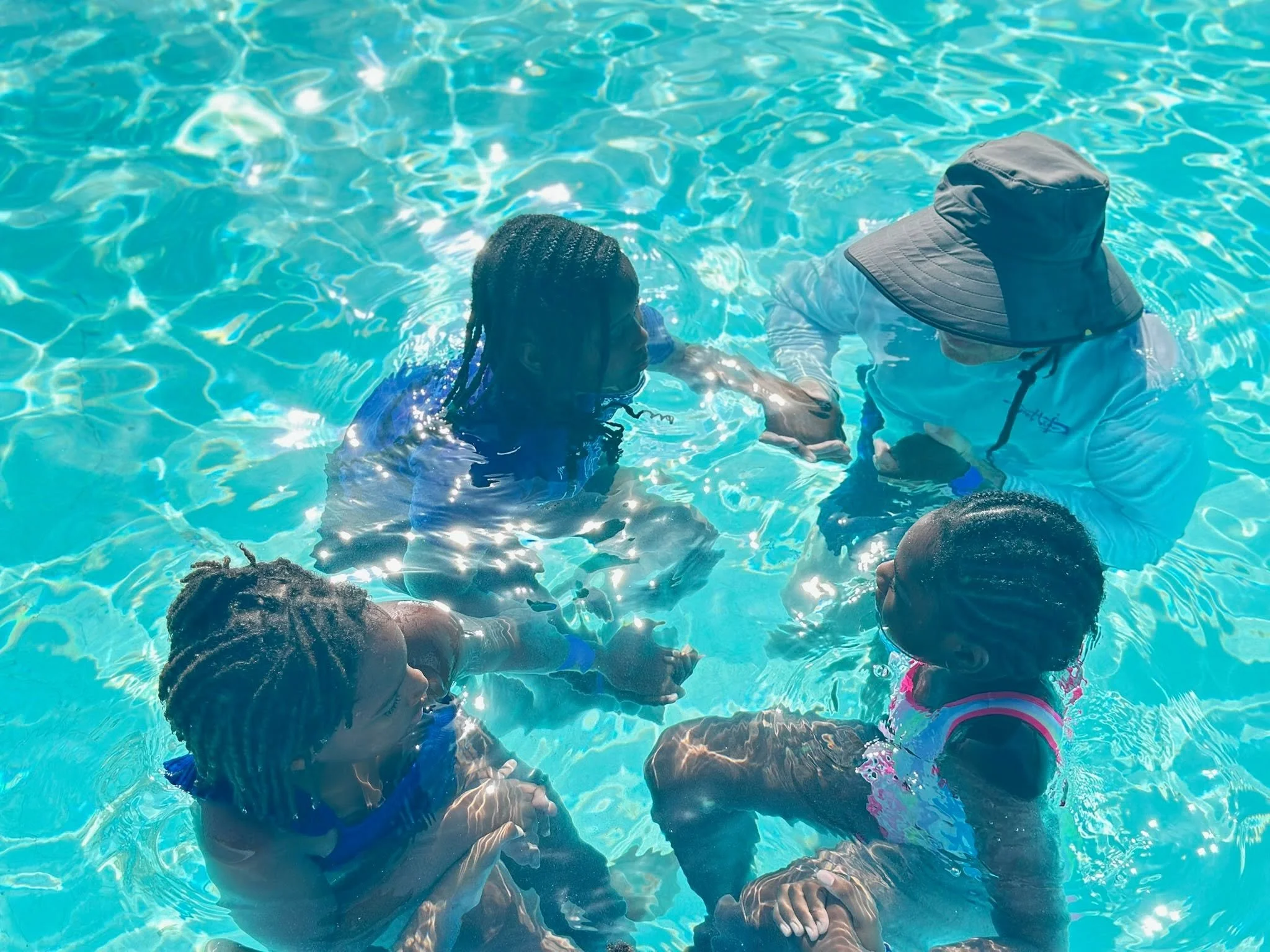 Children swimming in a pool with clear blue water, one girl with braids and others in colorful swimsuits.