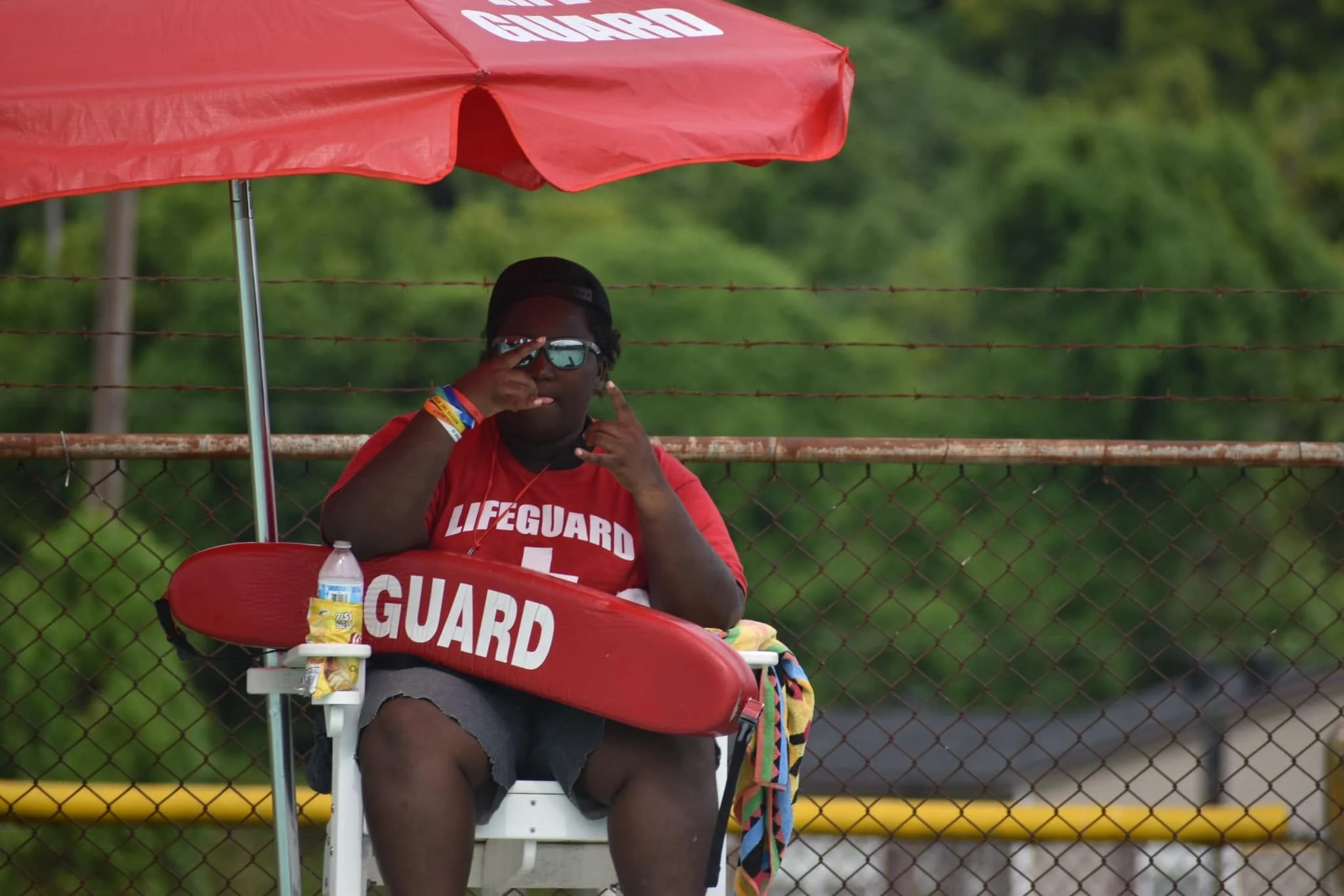 A lifeguard sitting on a chair under a red umbrella beside a fence, wearing sunglasses and a red shirt that says 'LIFEGUARD'. She is making a gesture with her hand near her face and has a water bottle and snacks on the chair.