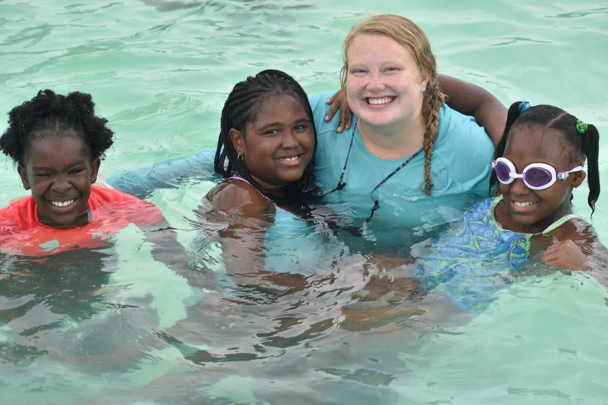 A group of four children and a young adult smiling and swimming in a pool, with two girls wearing goggles.