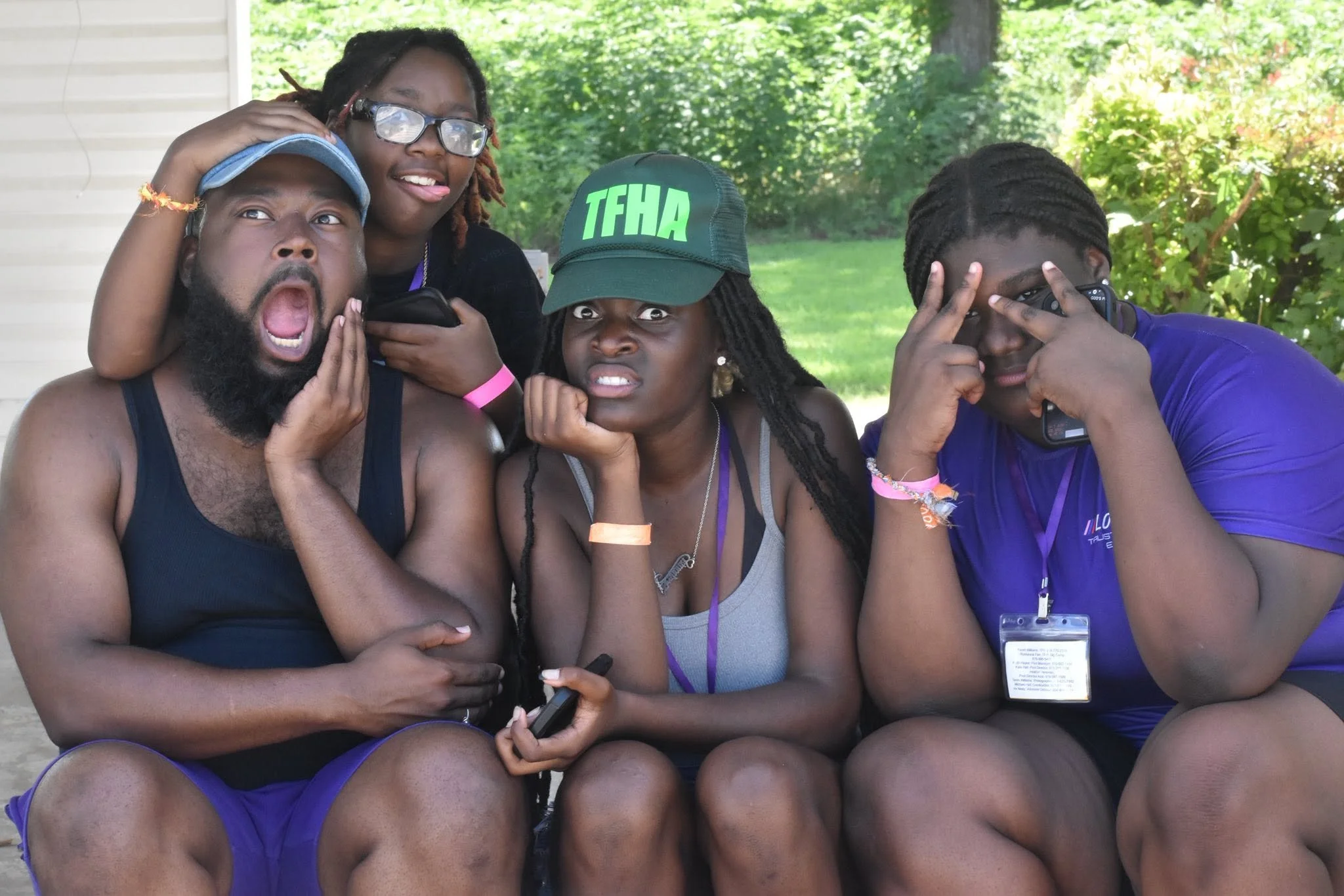 Group of five diverse young adults sitting outdoors, posing playfully for a photo. Some are holding phones, and one is wearing a cap with the acronym 'TFHA.' They are surrounded by greenery and appear to be having fun.