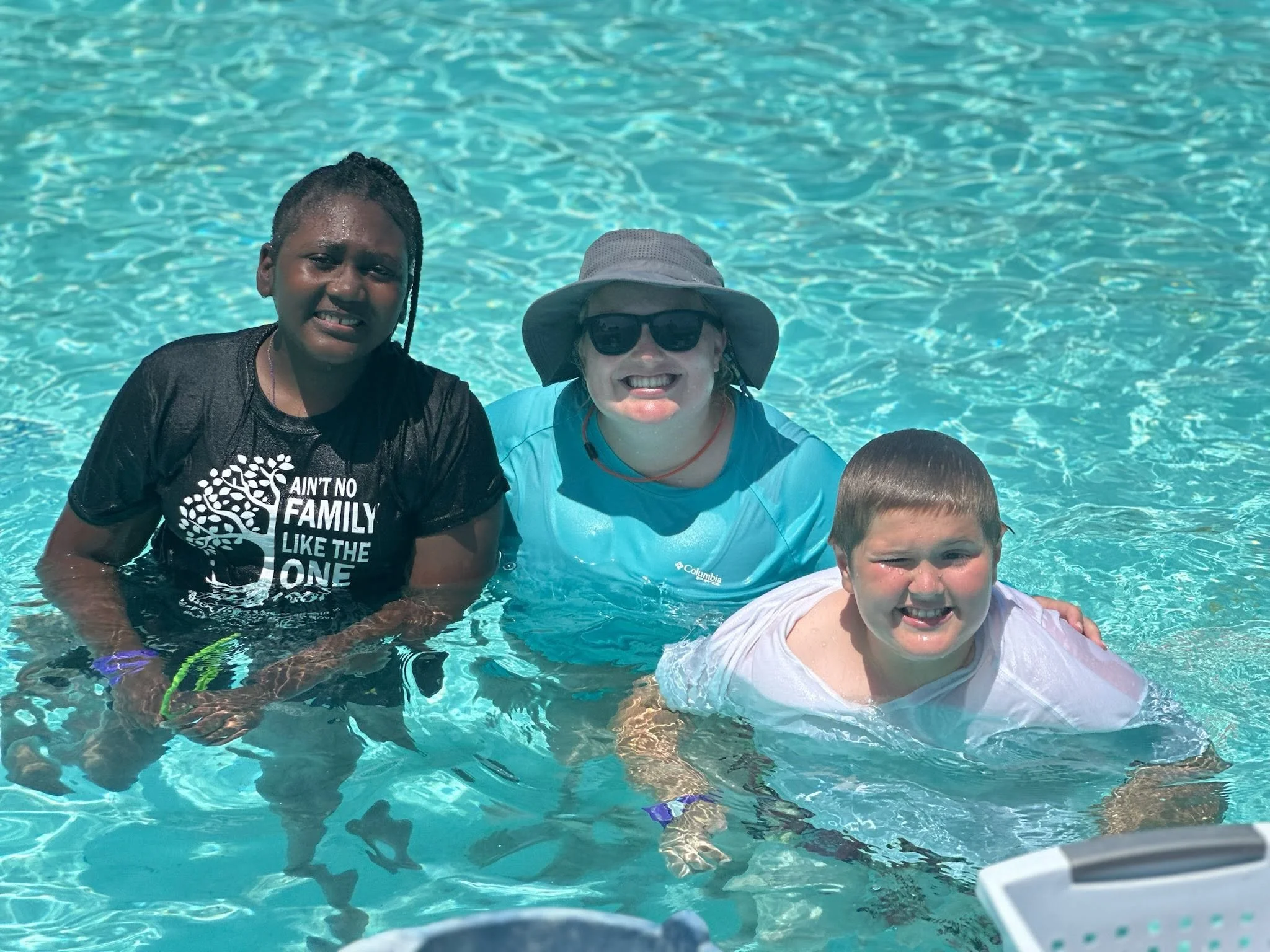 Three people posing in a swimming pool with water and their heads above the water, two are smiling and one appears to have a more serious expression, one person is wearing sunglasses and a sun hat.