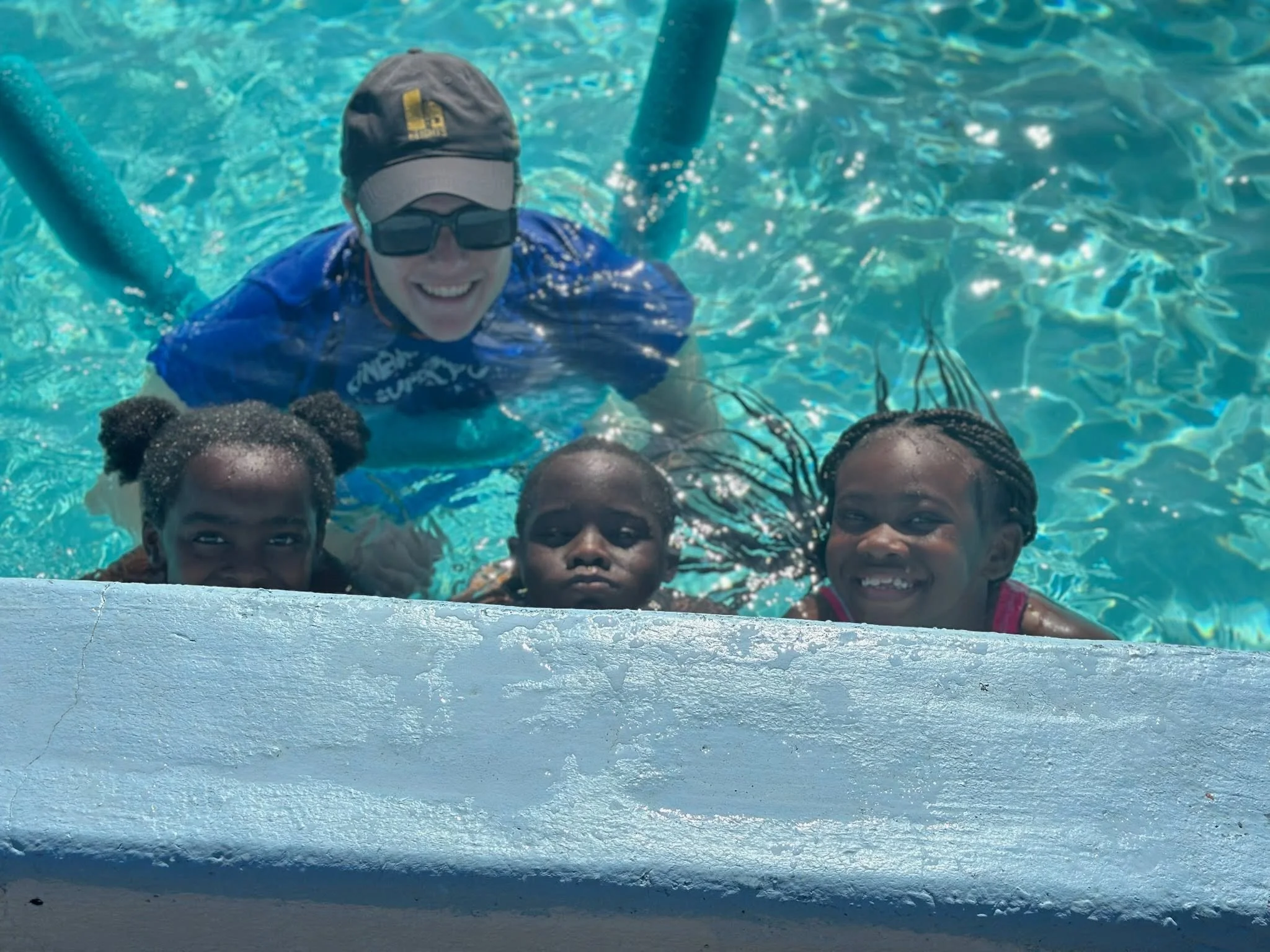 A group of children and a woman are in a swimming pool, with the woman smiling and wearing sunglasses and a cap. The children are mostly submerged, with their heads above the water, and appear to be happy.