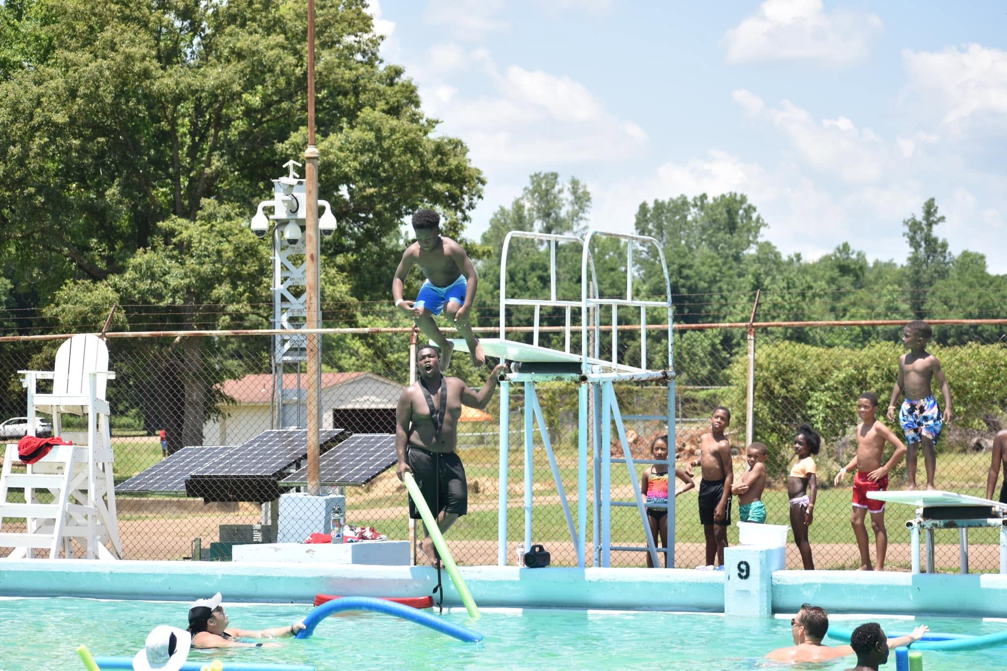 Children and teenagers at a swimming pool with a diving board, some preparing to jump and others standing nearby, surrounded by safety fencing and trees.