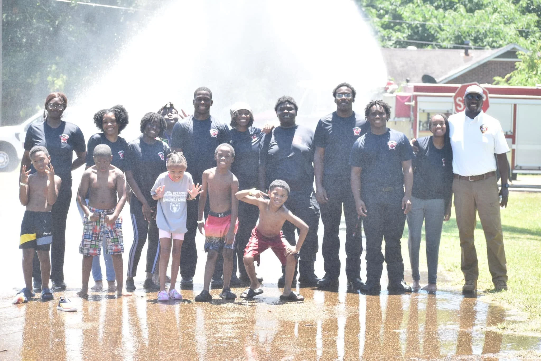 Group of children and adults standing in front of a water spray, smiling, outdoors on a sunny day, some children are shirtless, some wearing swim trunks, and some wearing t-shirts with team logos.