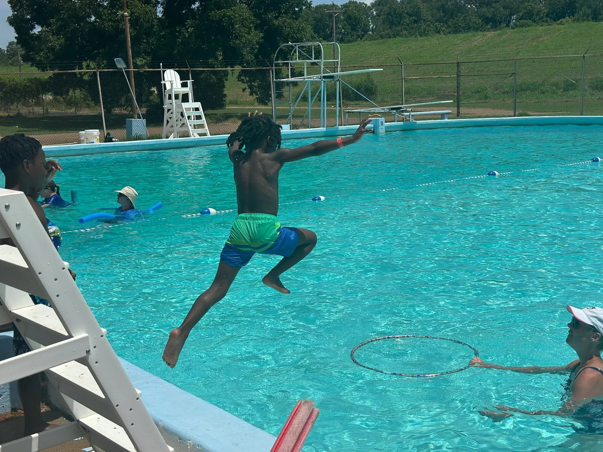 Children playing and jumping into a swimming pool, with two adults supervising nearby, surrounded by a chain-link fence and grassy area.