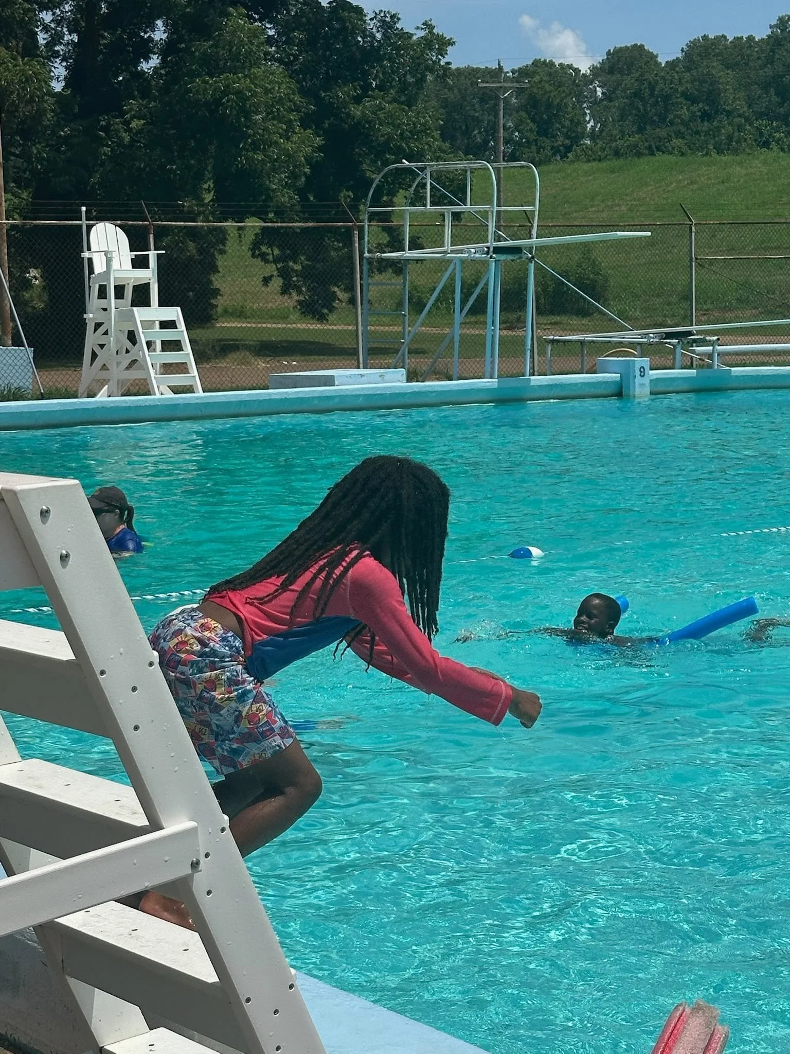 A swimming pool with two children playing in the water, one girl with braided hair wearing a pink long-sleeve shirt and colorful shorts, and a boy floating with a blue foam noodle. An adult is sitting at the edge of the pool, partly visible. There is