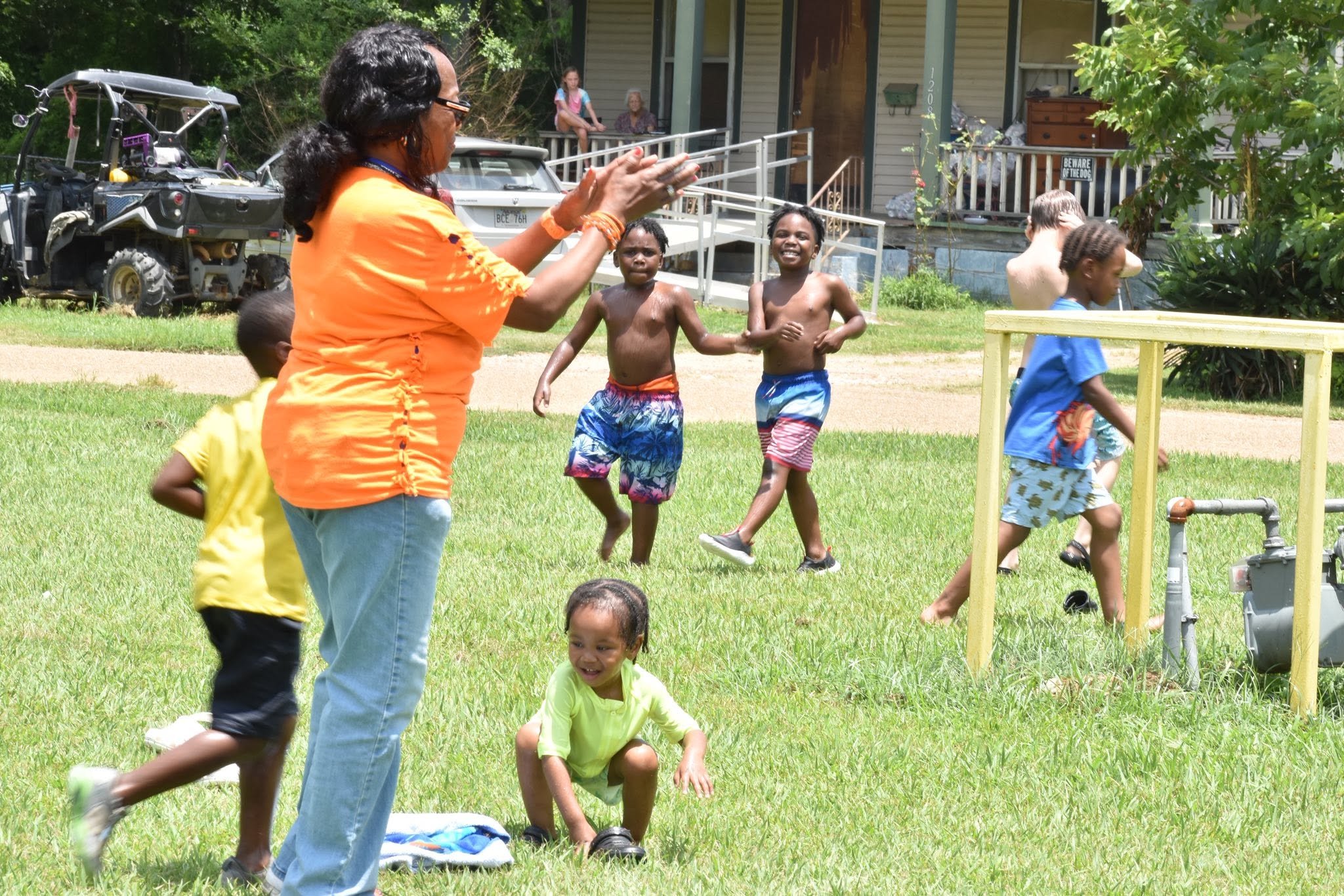 A woman clapping and smiling while children play outside on the grass near a house and a wheelchair ramp.