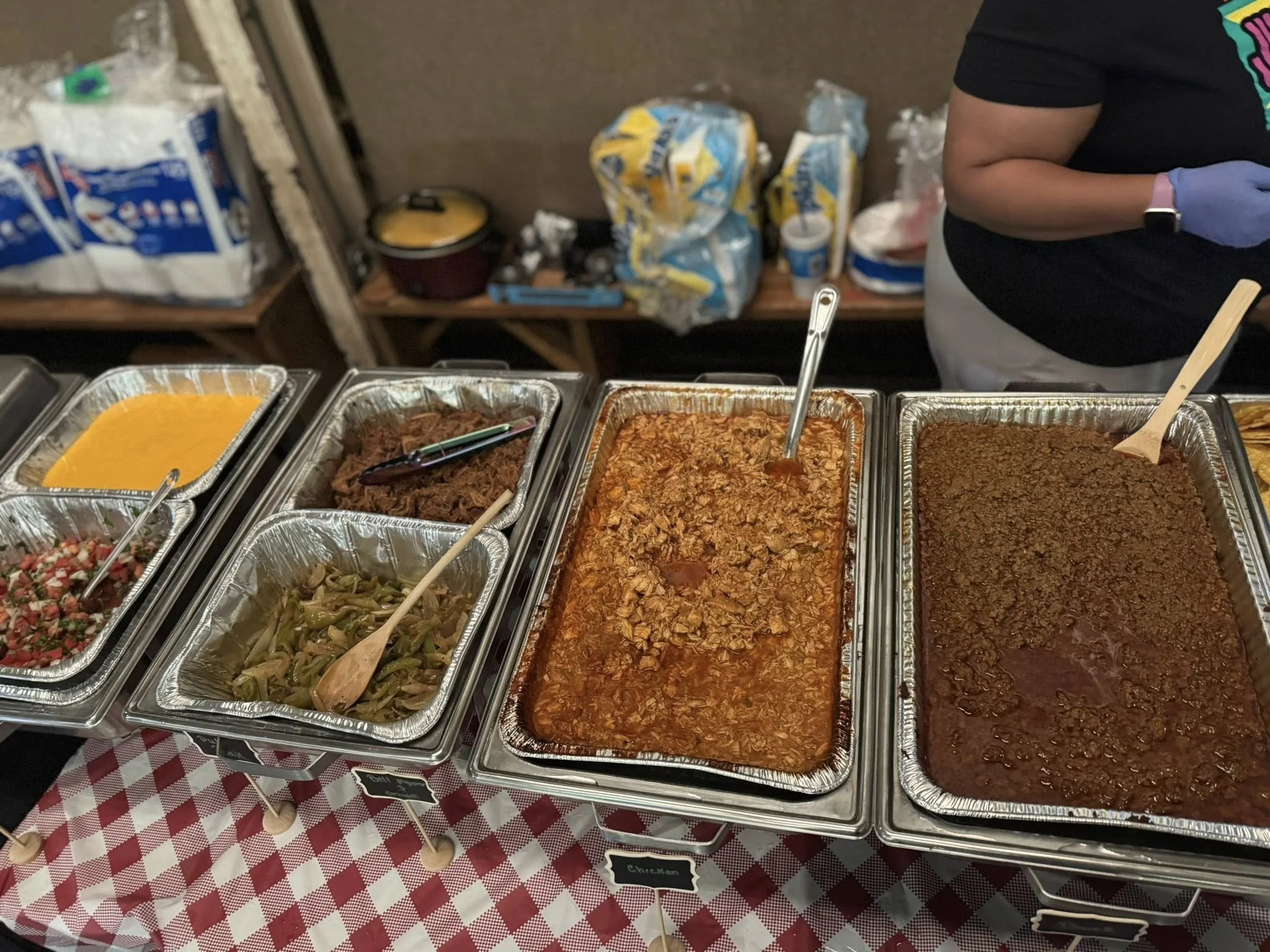 A buffet with six aluminum trays of food, including mashed sweet potatoes, shredded beef, barbecue chicken, and ground beef, on a table with a red and white checkered tablecloth.