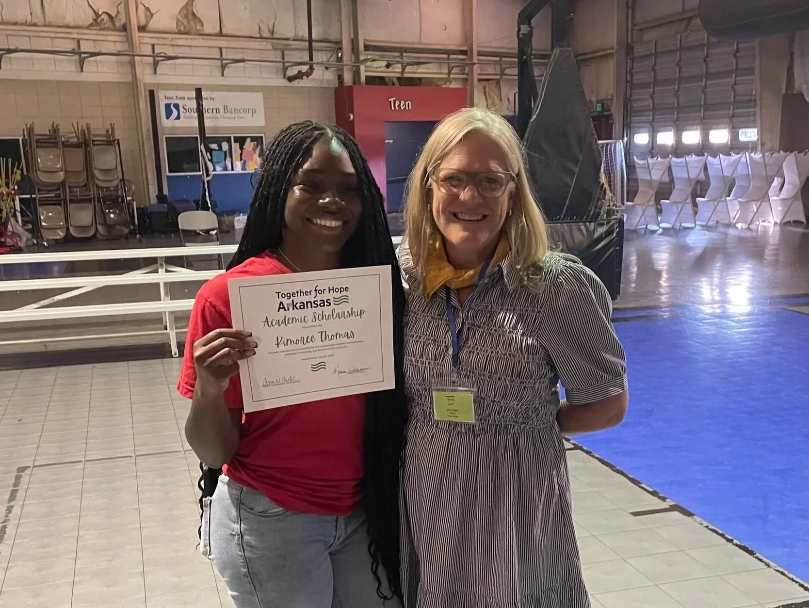 A young woman holding a certificate that reads 'Together for Hope Arkansas Academic Scholarship Kimalee Thomas,' standing next to an older woman in a striped dress and glasses inside a gymnasium or event hall with chairs and stage setup in the background.