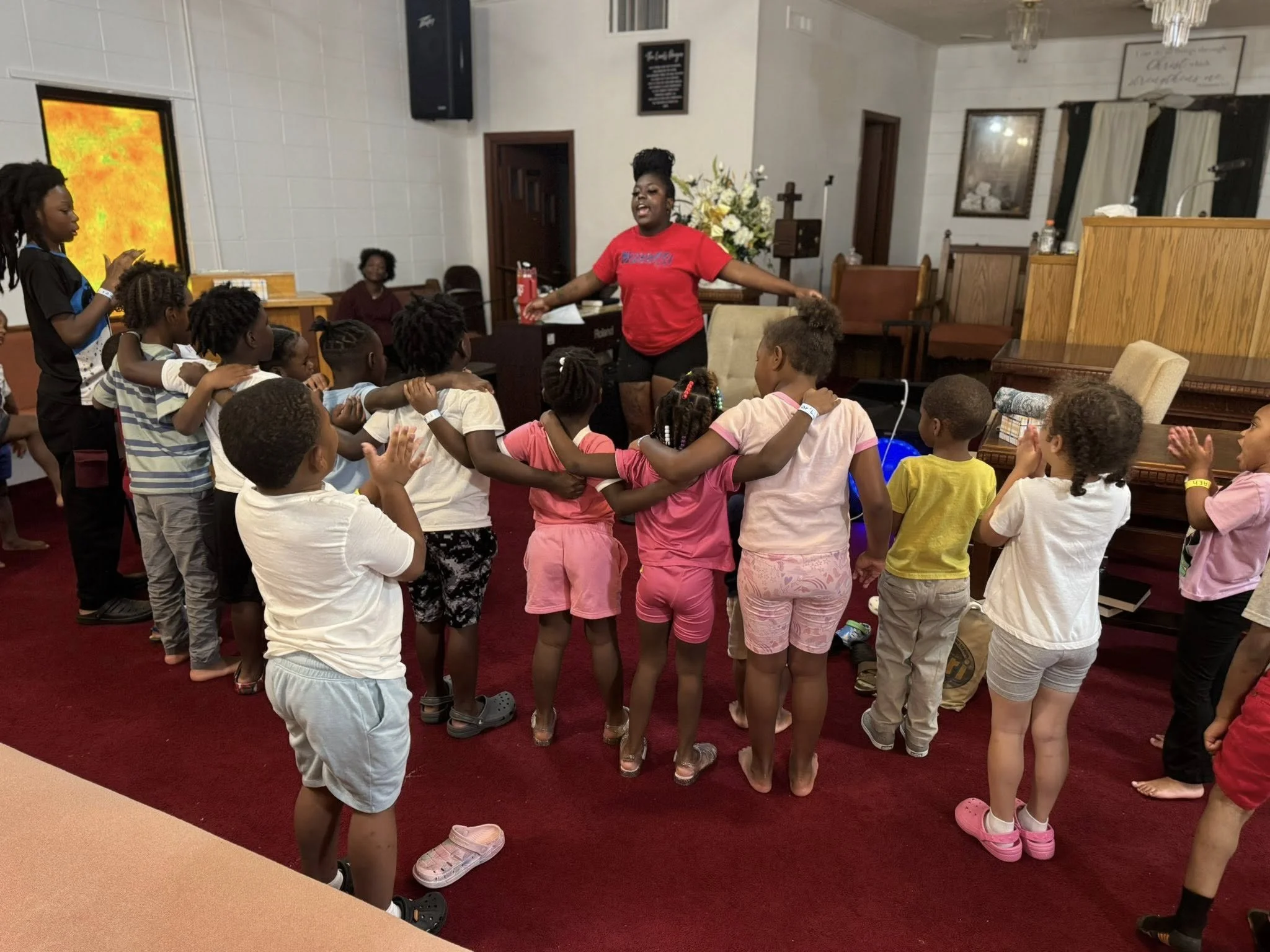 A group of children gathered in a church, watching a woman in a red shirt dance or perform in front of them. Some children are standing, others sitting, and all appear engaged. The church has white walls, wooden furniture, and a floral arrangement in