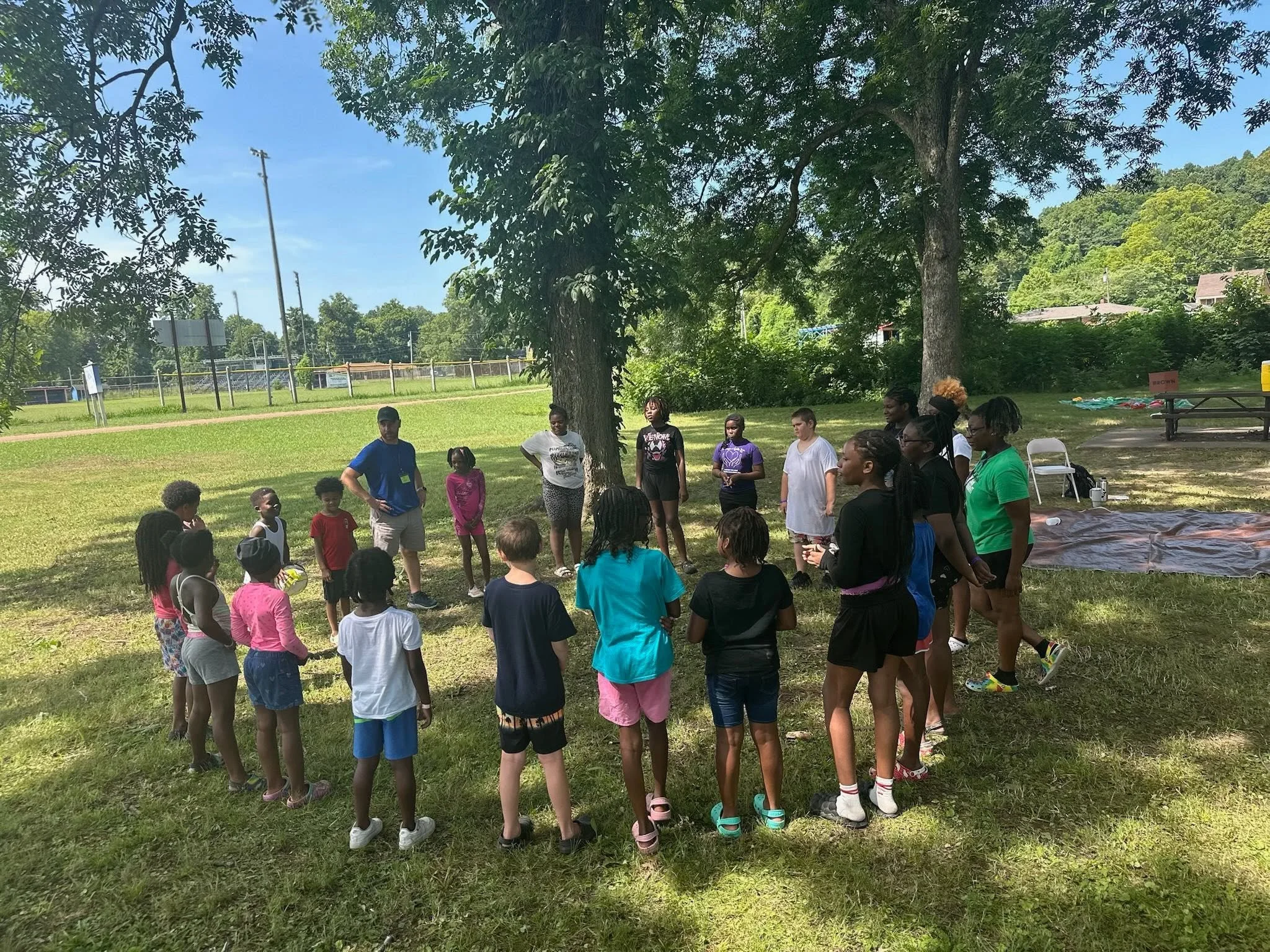 A diverse group of children and adults gathered outdoors under trees in a park-like setting, participating in an event or activity.