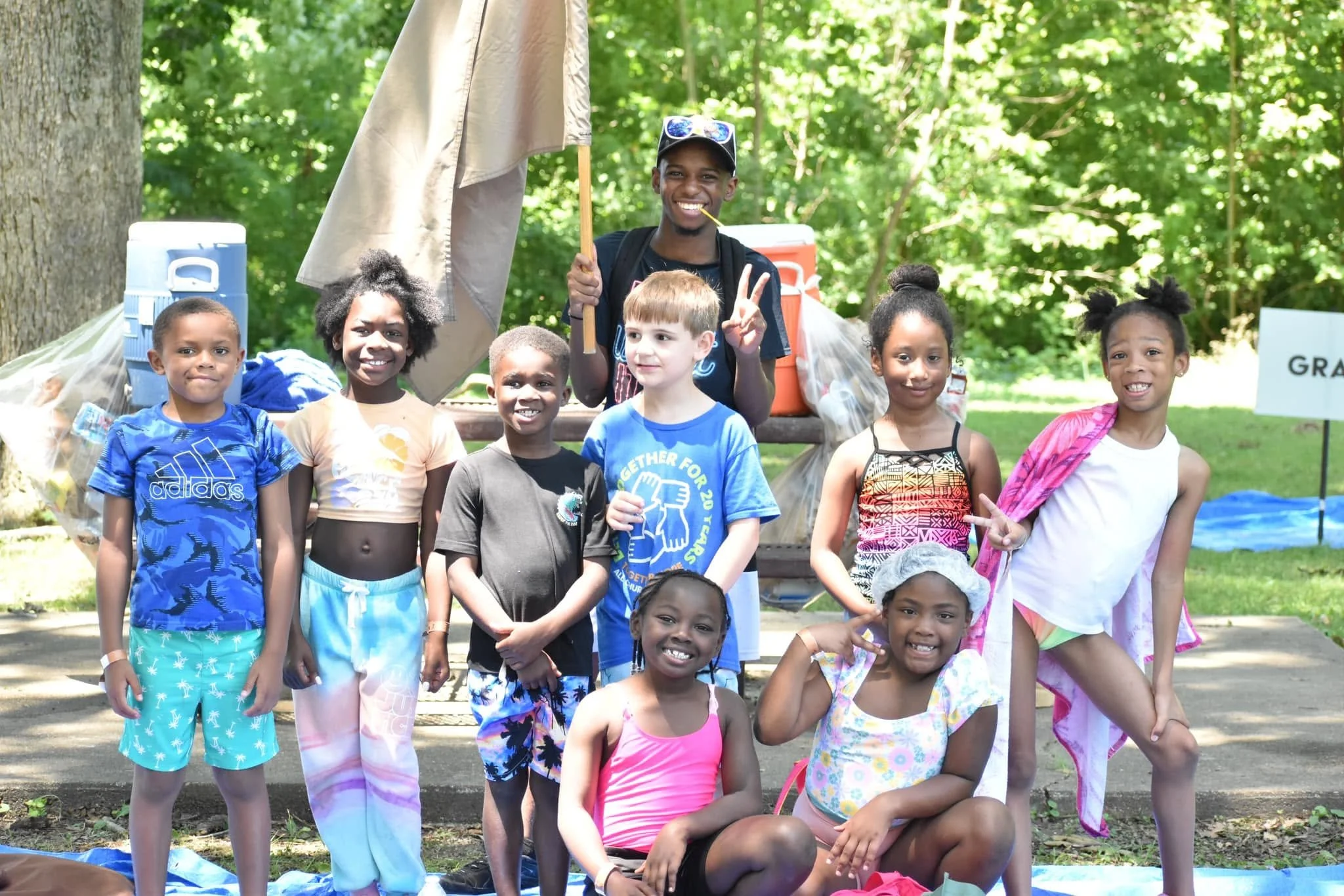 A group of children and a young man outdoors in a wooded area posing for a photo. The young man is smiling and holding a peace sign, wearing sunglasses and a cap. The children are dressed in colorful casual clothing and are smiling.