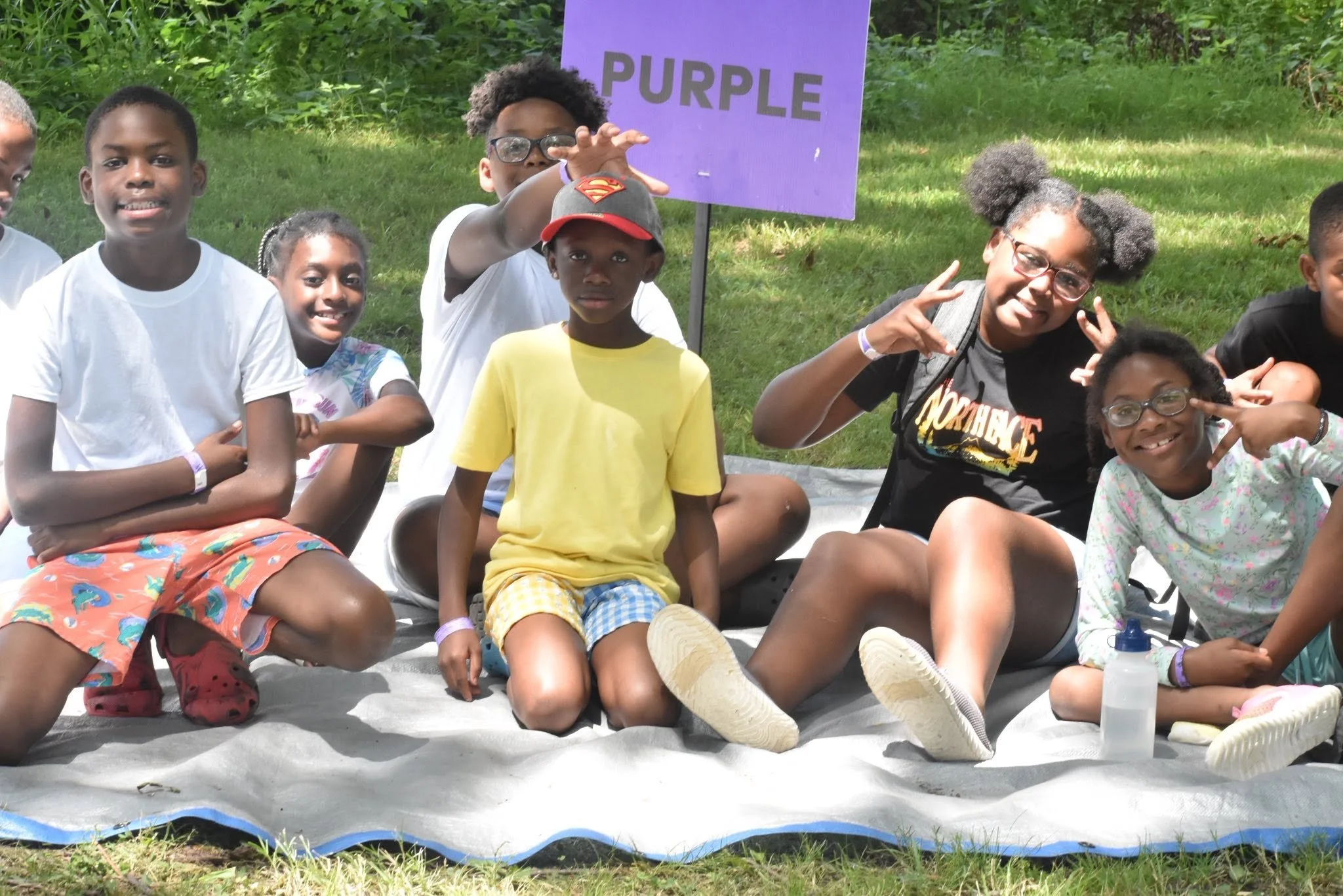 A group of children sitting on a blanket outdoors with a purple sign that says 'PURPLE' behind them. They are smiling and making playful hand gestures.