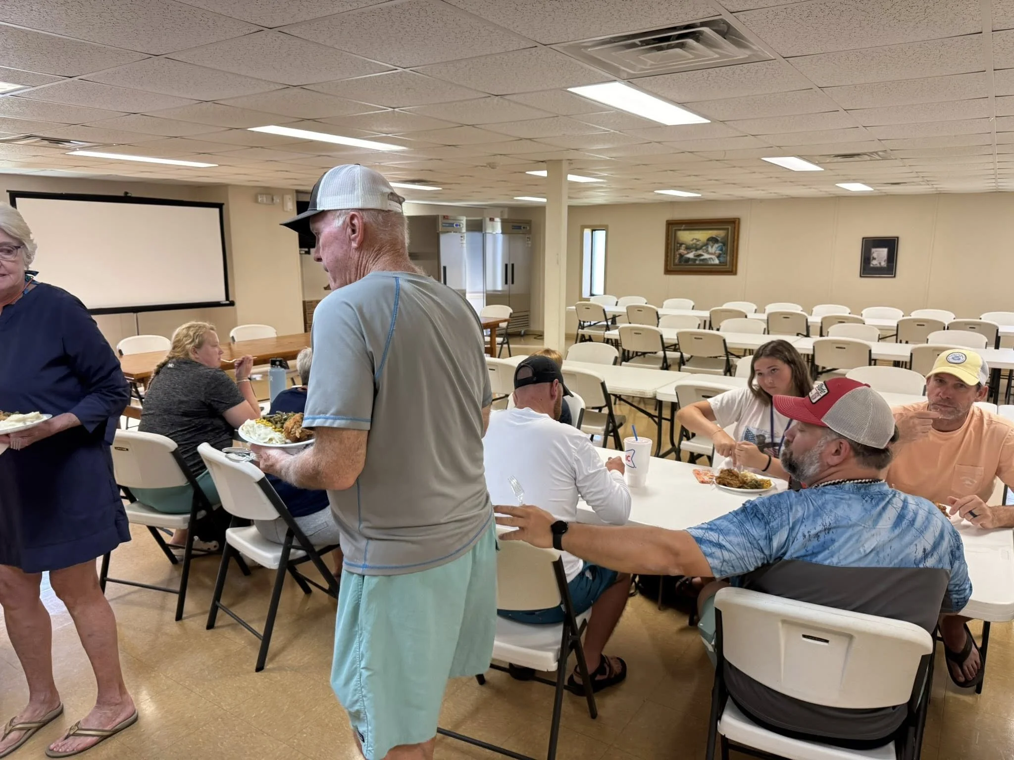 People gathered in a room with folding chairs and tables, some eating food, others standing and talking, with paintings on the wall and a projection screen in the background.