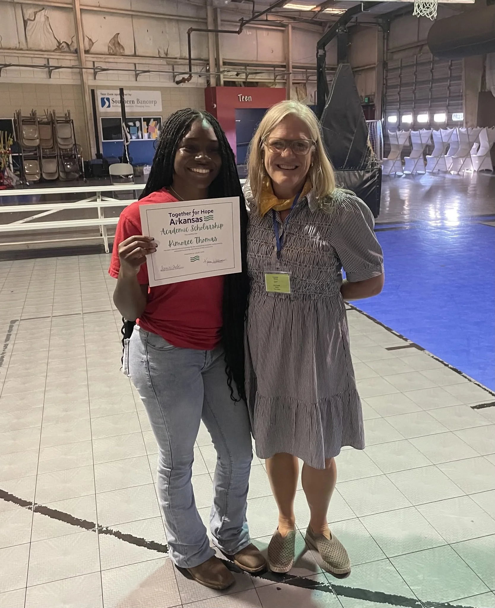 Two women standing inside a gymnasium, one holding a certificate for an academic scholarship from Arkansas, and both smiling at the camera. The woman on the left is wearing a red top and light blue jeans, and the woman on the right is wearing glasses