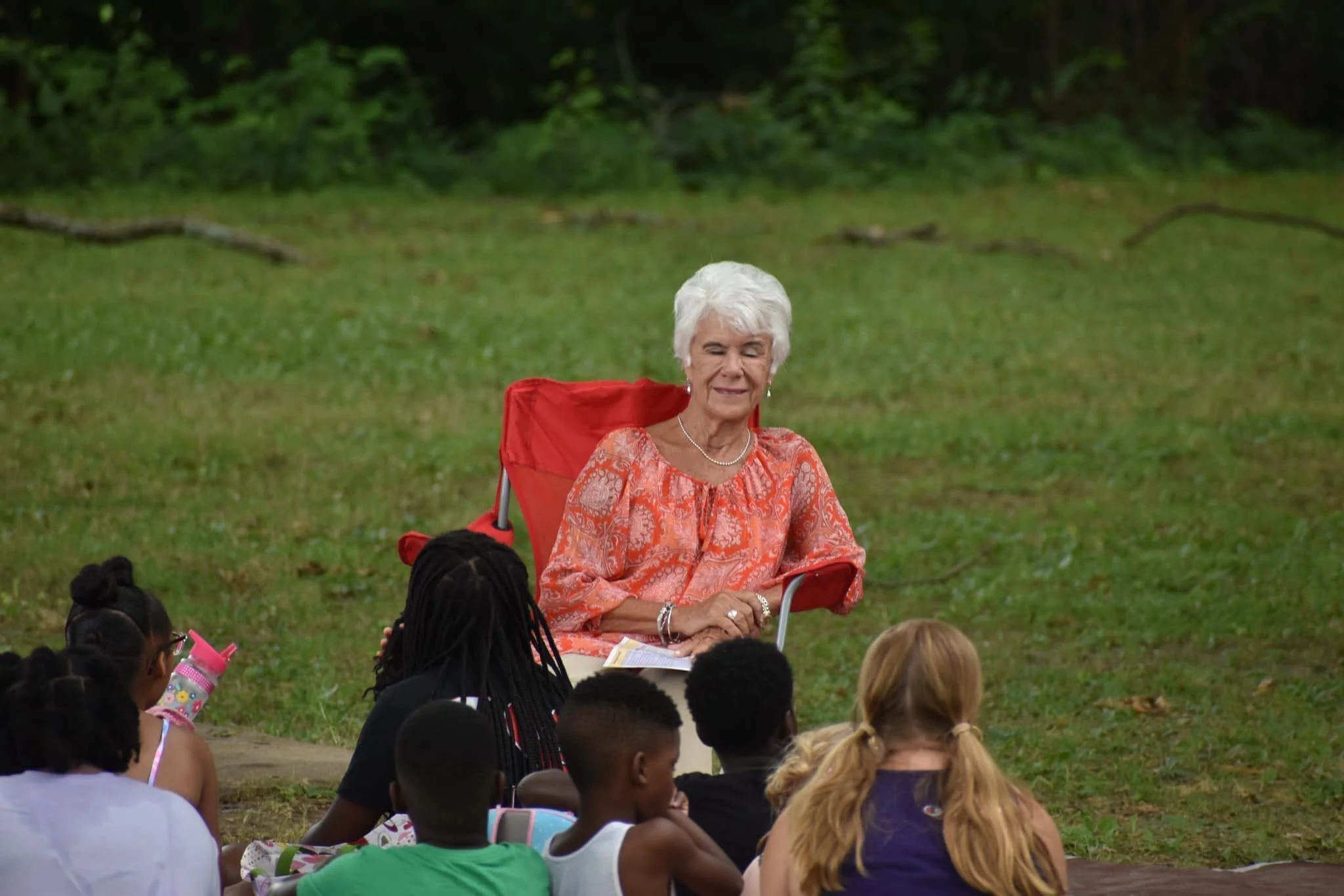 An elderly woman with white hair and earrings, sitting on a red folding chair outdoors on grass, surrounded by a group of children, with a wooded area in the background.