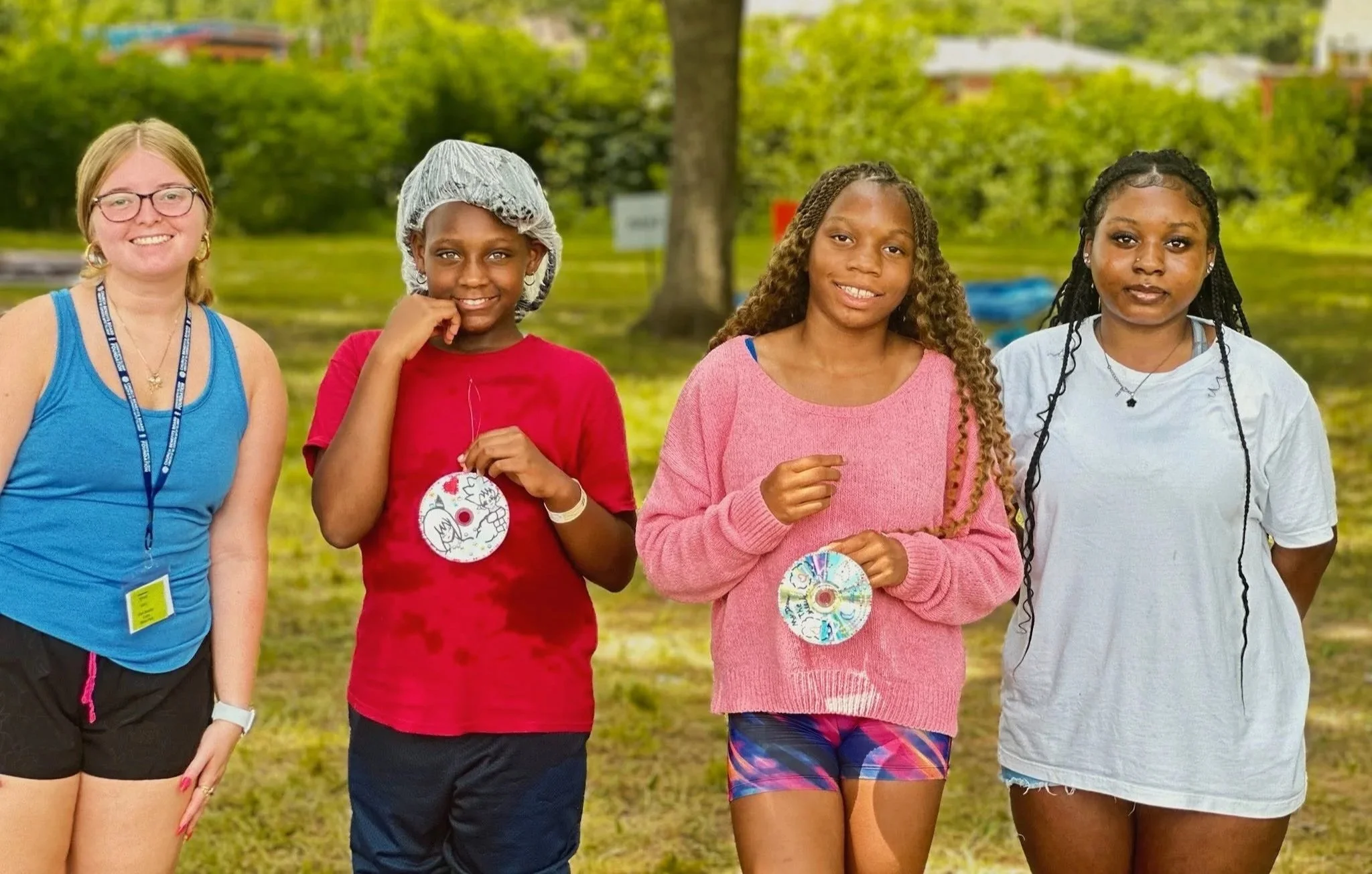 Four women standing outdoors in a park, smiling for the camera. One woman is wearing a blue tank top, another in a red shirt with a hairnet, a third woman in a pink sweater and colorful shorts, and the fourth in a white t-shirt with braided hair.