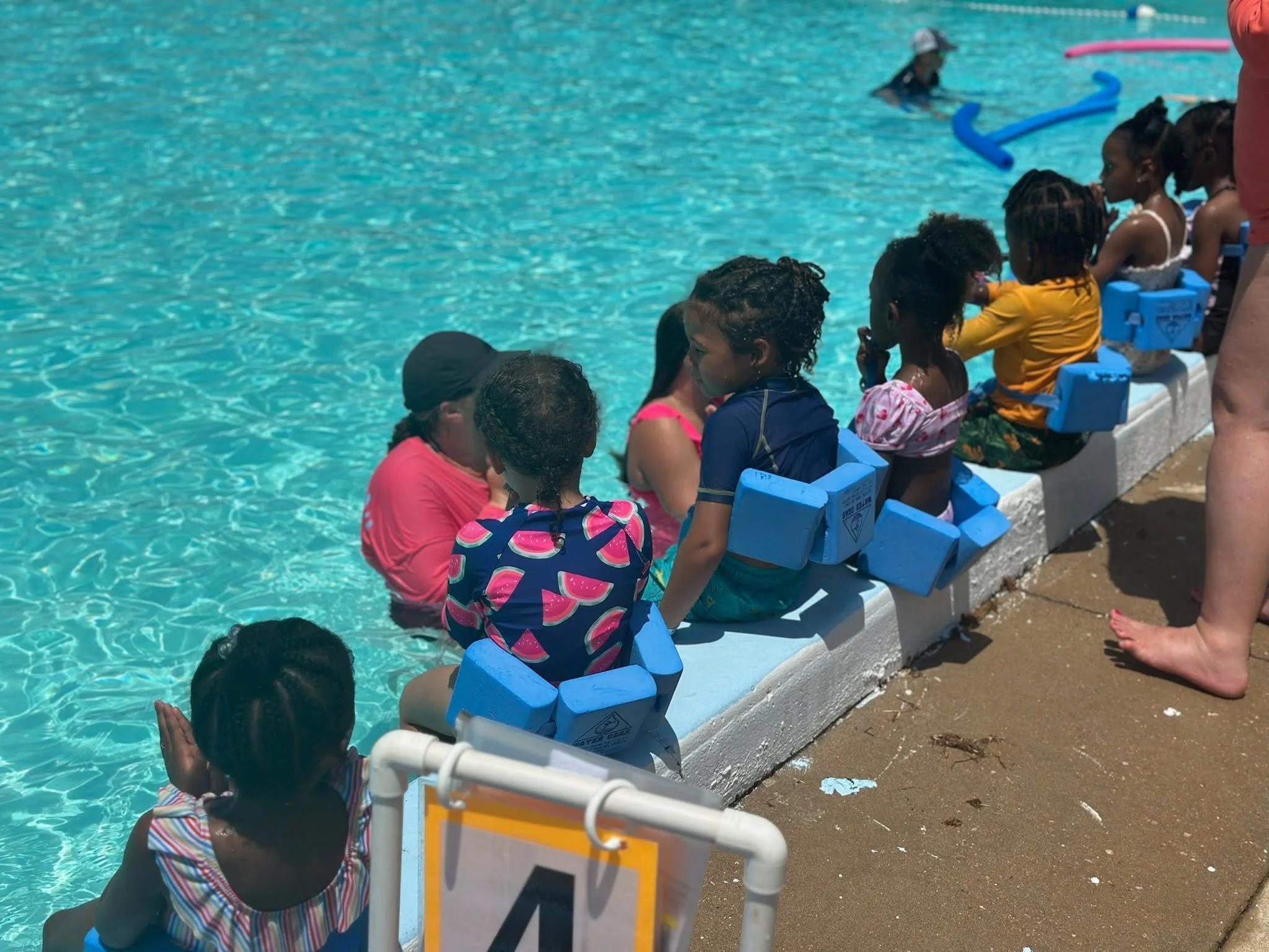 A group of children sitting on blue chairs by a swimming pool, with some children in the water and others waiting on the poolside, engaging in a swimming lesson or activity.