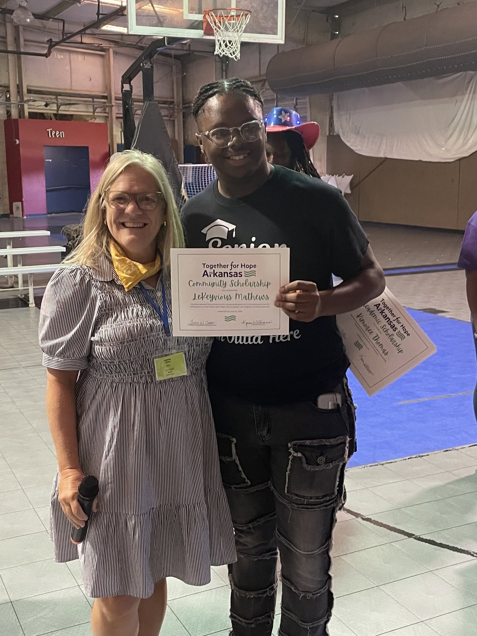A woman with blond hair, glasses, and a striped dress holding a microphone stands next to a smiling young man with glasses, braided hair, wearing a black T-shirt and cargo shorts. He is holding a certificate that reads 'Together for Hope Arkansas Community Scholarship Lakeview Thomas'. They are in a gymnasium with a basketball hoop in the background.