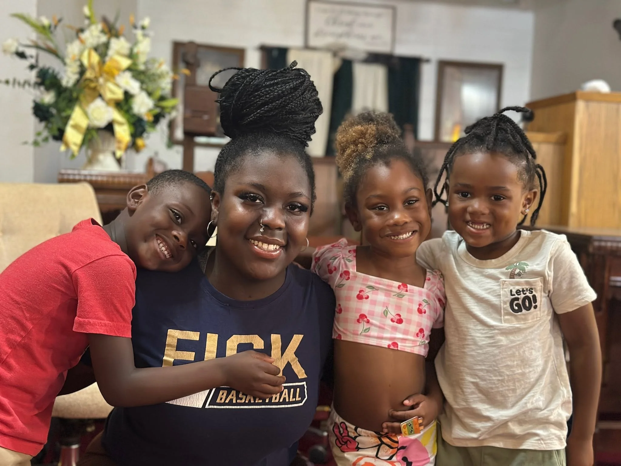 A group of four children and one adult girl smiling and posing together indoors, with a floral arrangement and framed pictures visible in the background.