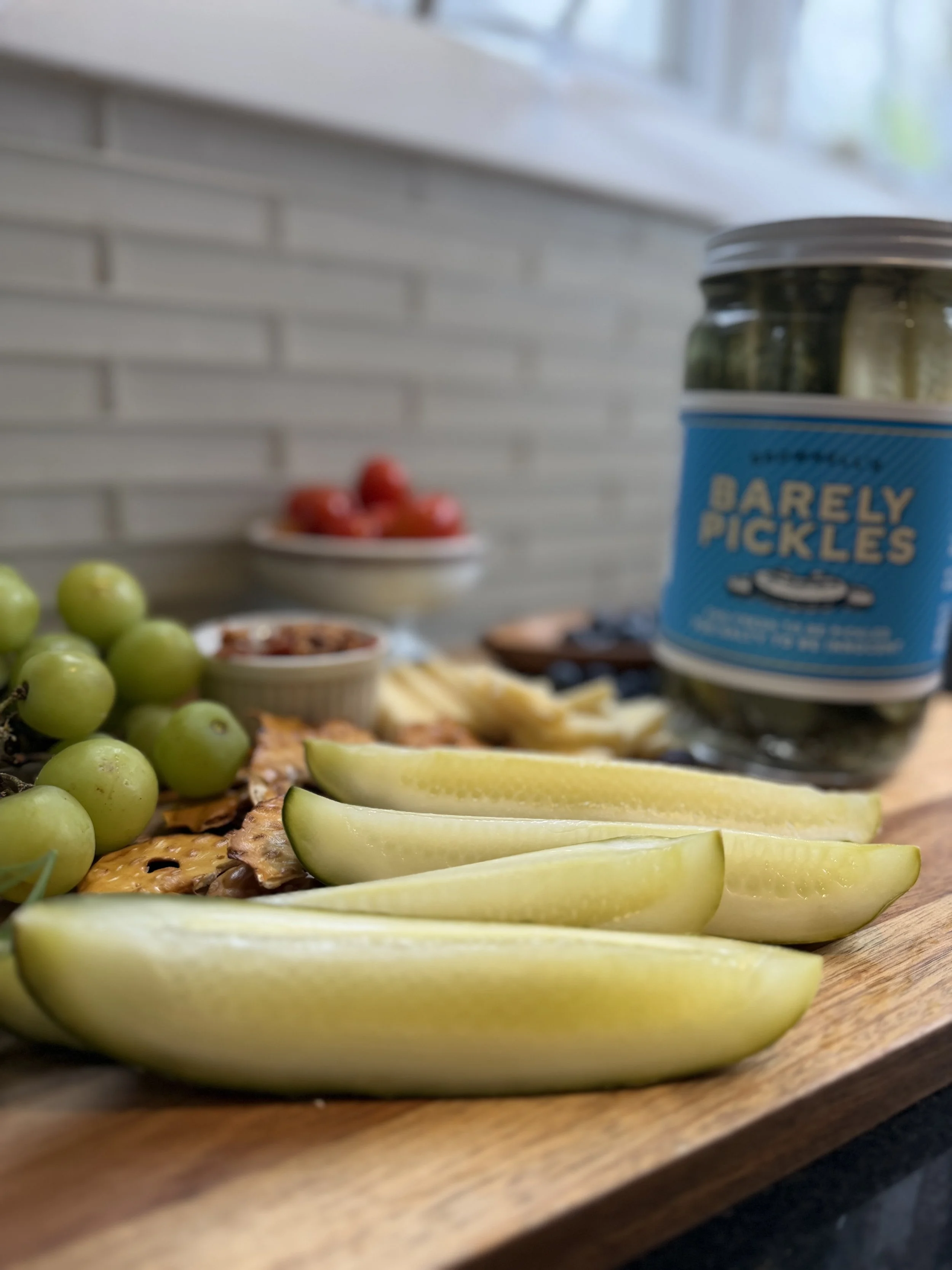 Close-up view of sliced yellow honeydew melon, green grapes, and crackers on a wooden serving board with a jar of dill pickles in the background, on a tabletop with a white brick wall and window in the background.