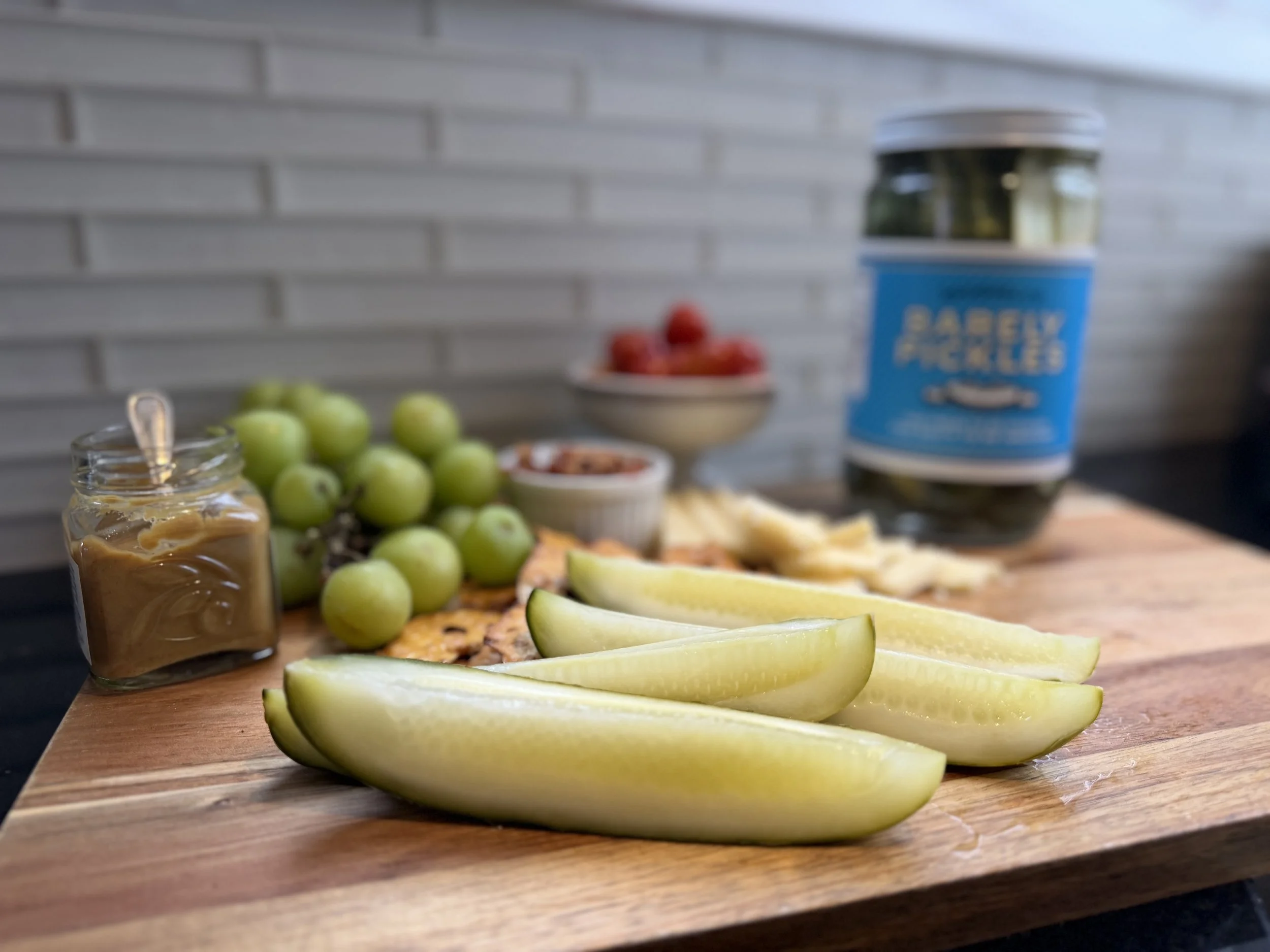 A cutting board with sliced yellow pears in the foreground, a jar of caramel, green grapes, cherry tomatoes in a small bowl, and slices of lemon. In the background, a container labeled 'Baby Snacks' and a bowl of cherry tomatoes are visible. The backdrop is a white brick wall.