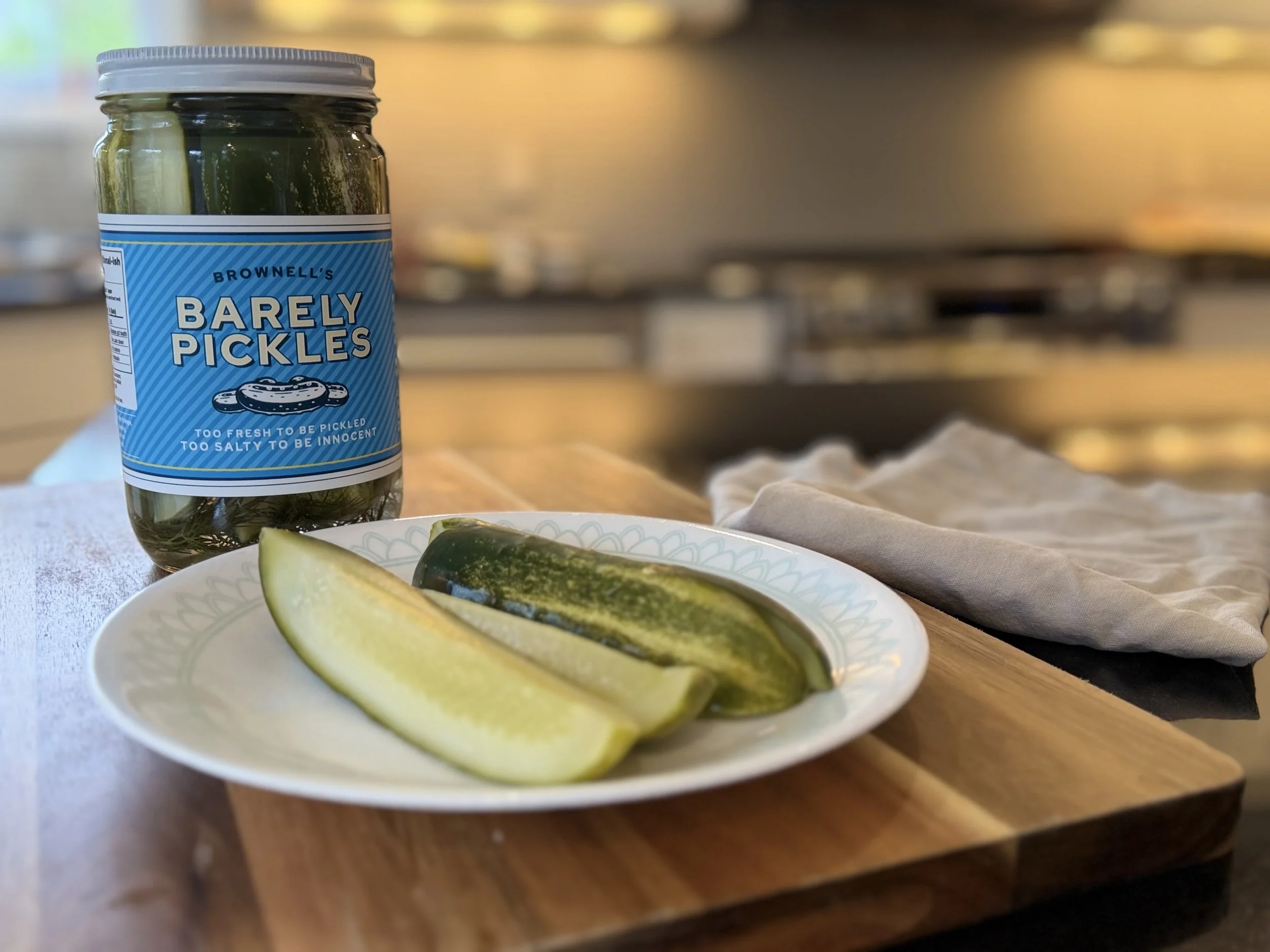 A jar labeled 'Barely Pickles' on a wooden table, with sliced cucumber on a plate and a gray cloth napkin nearby. The background is blurred.