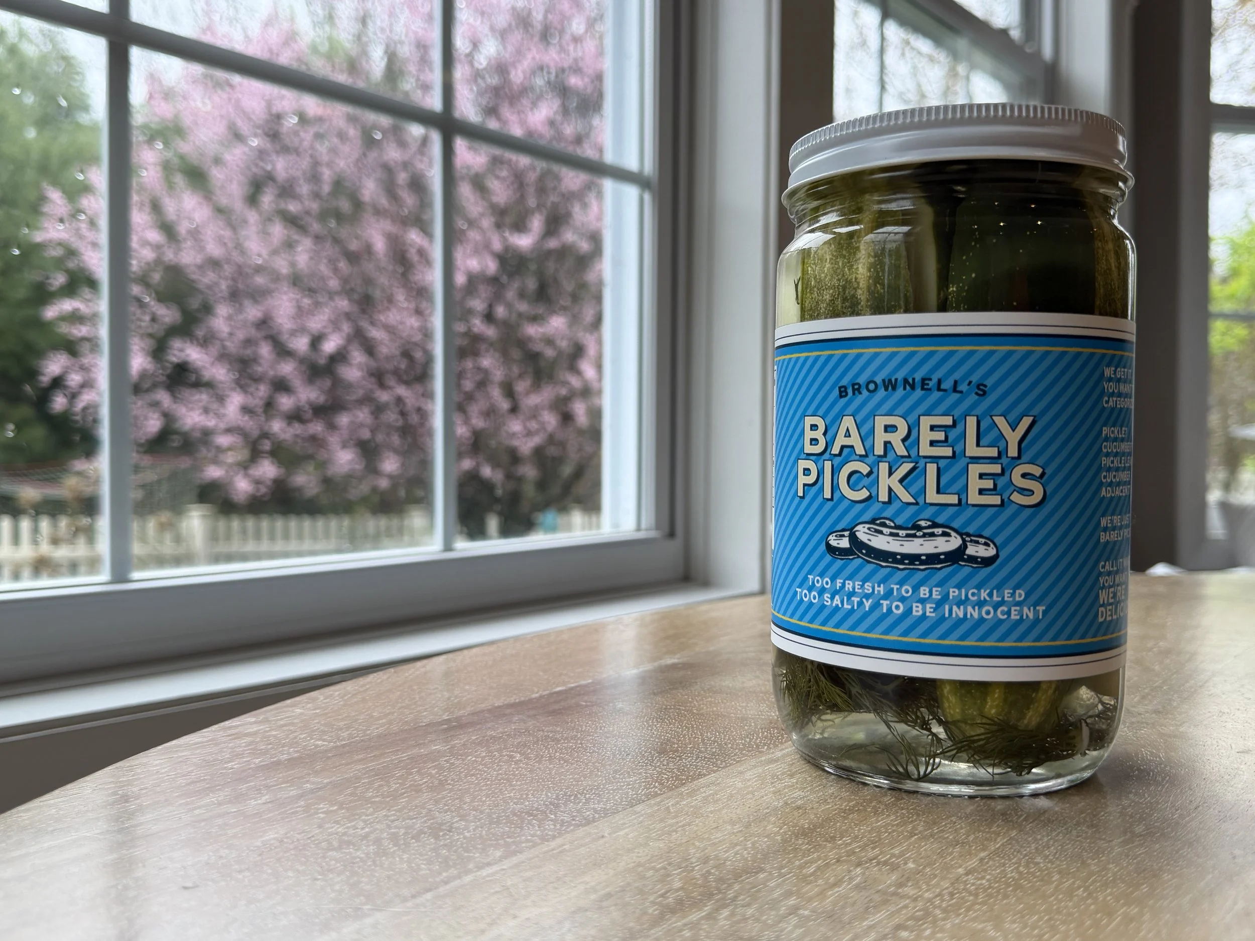 A jar of Brawell's Barely Pickles placed on a countertop by a window with pink and green foliage visible outside.